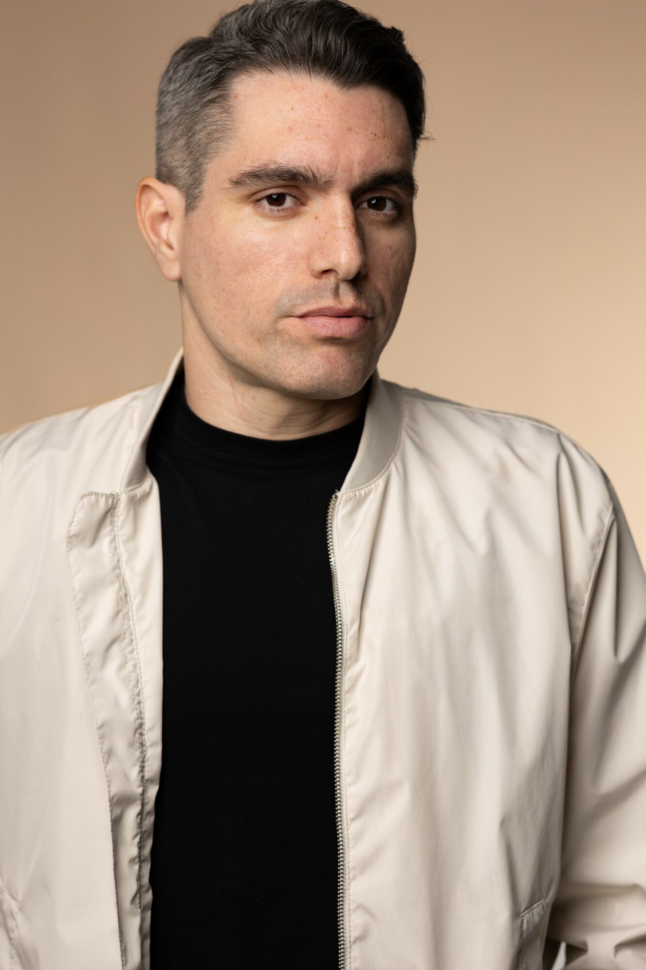 Man with short dark hair, wearing a black shirt and light-colored jacket, looking at the camera against a beige background.