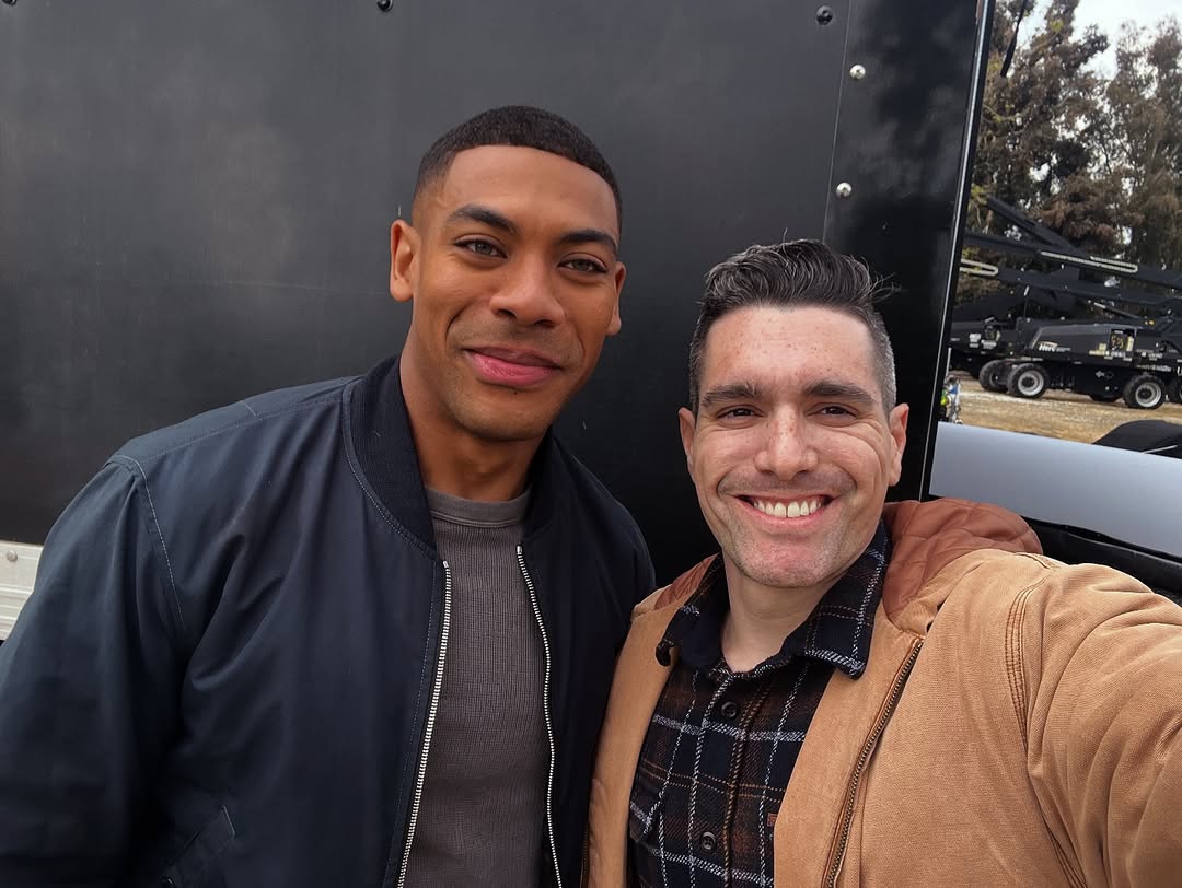 Two men smiling, one wearing a black jacket and the other a brown jacket, standing outdoors near a black background.