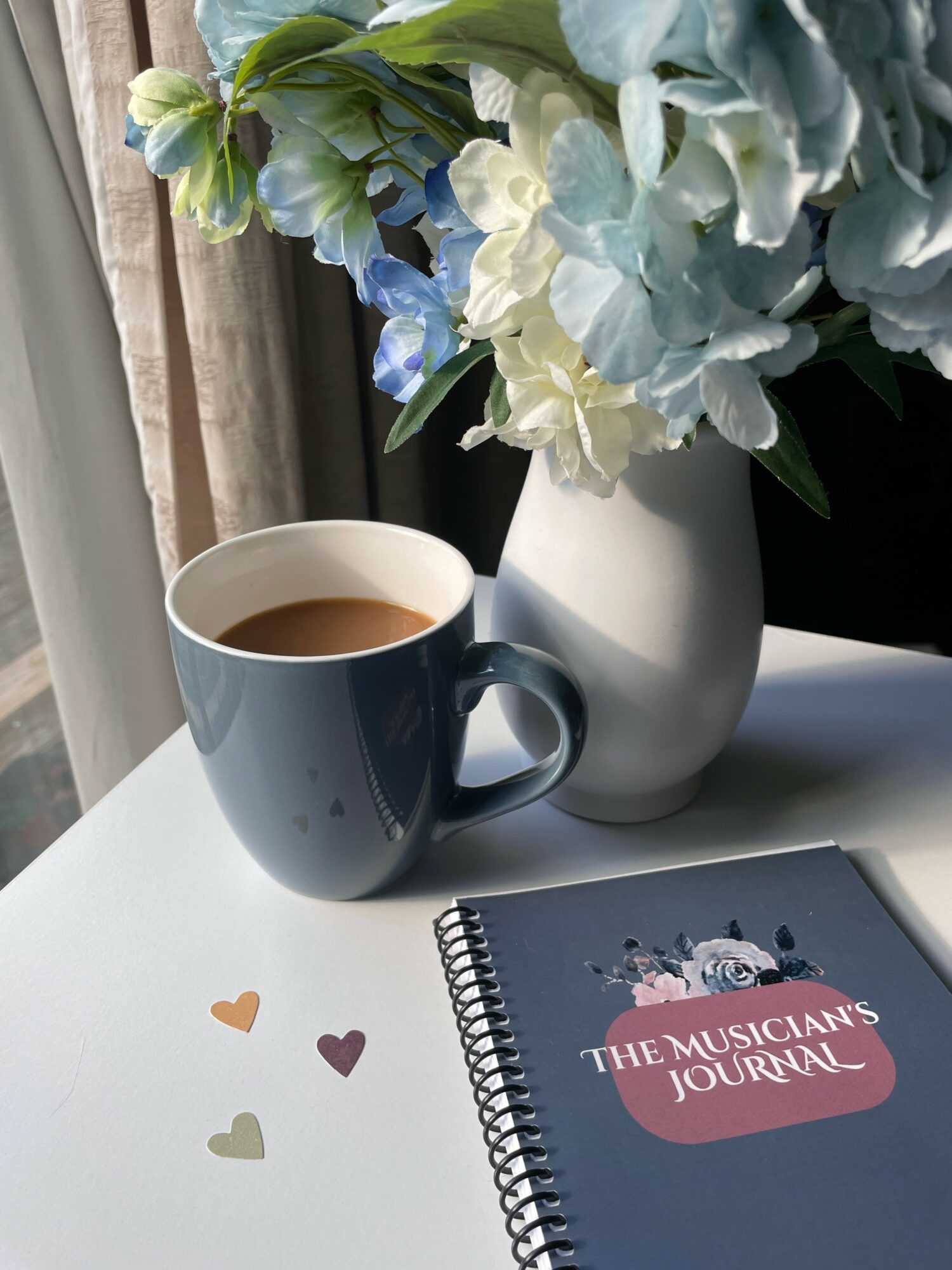 A blue mug filled with tea, a white vase with blue and white hydrangea flowers, a notebook titled 'The Musician's Journal', and small heart-shaped stickers on a white surface.