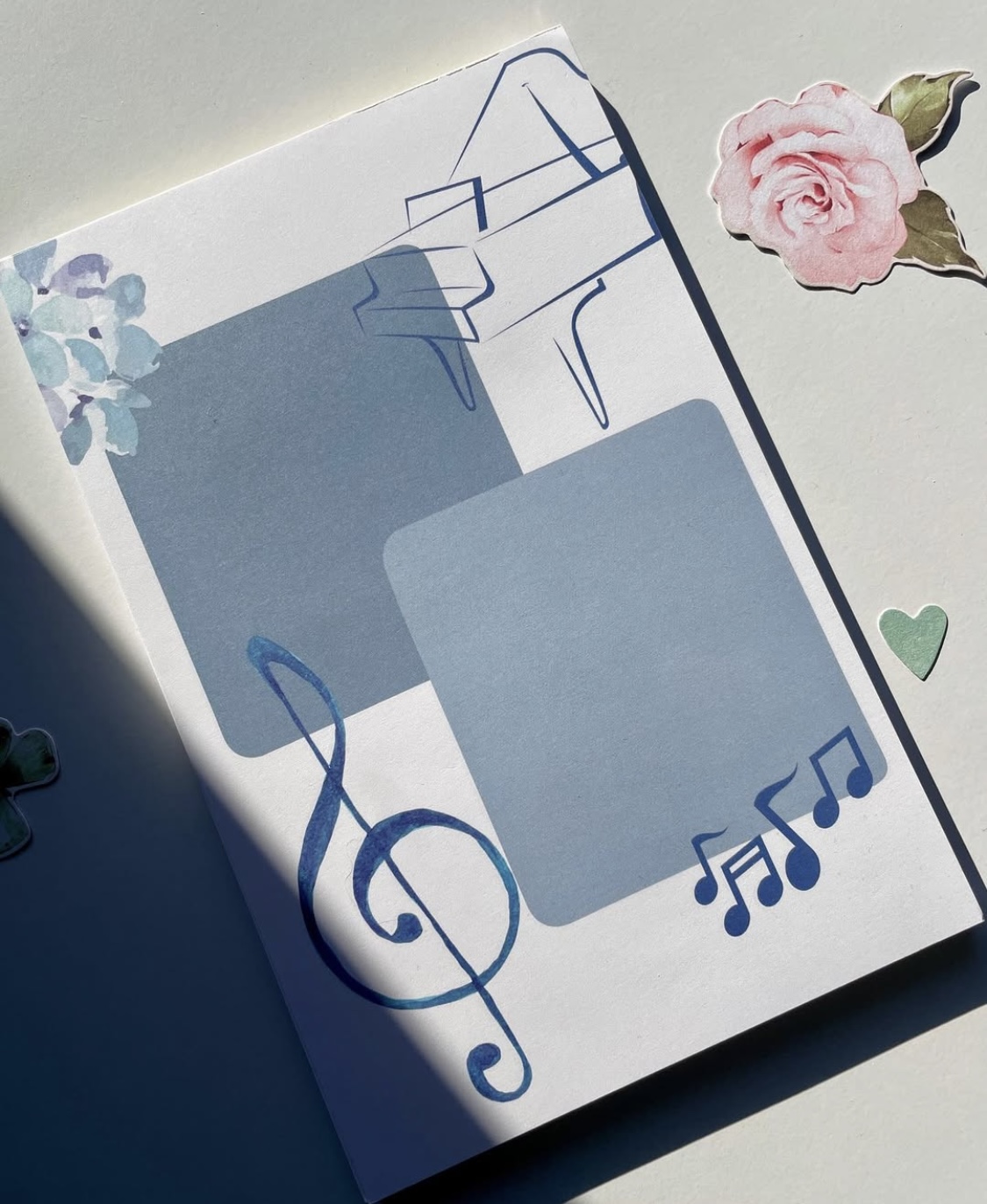 Open notebook with blue and white pages, decorated with a pink rose, green heart, and musical notes, on a light surface.