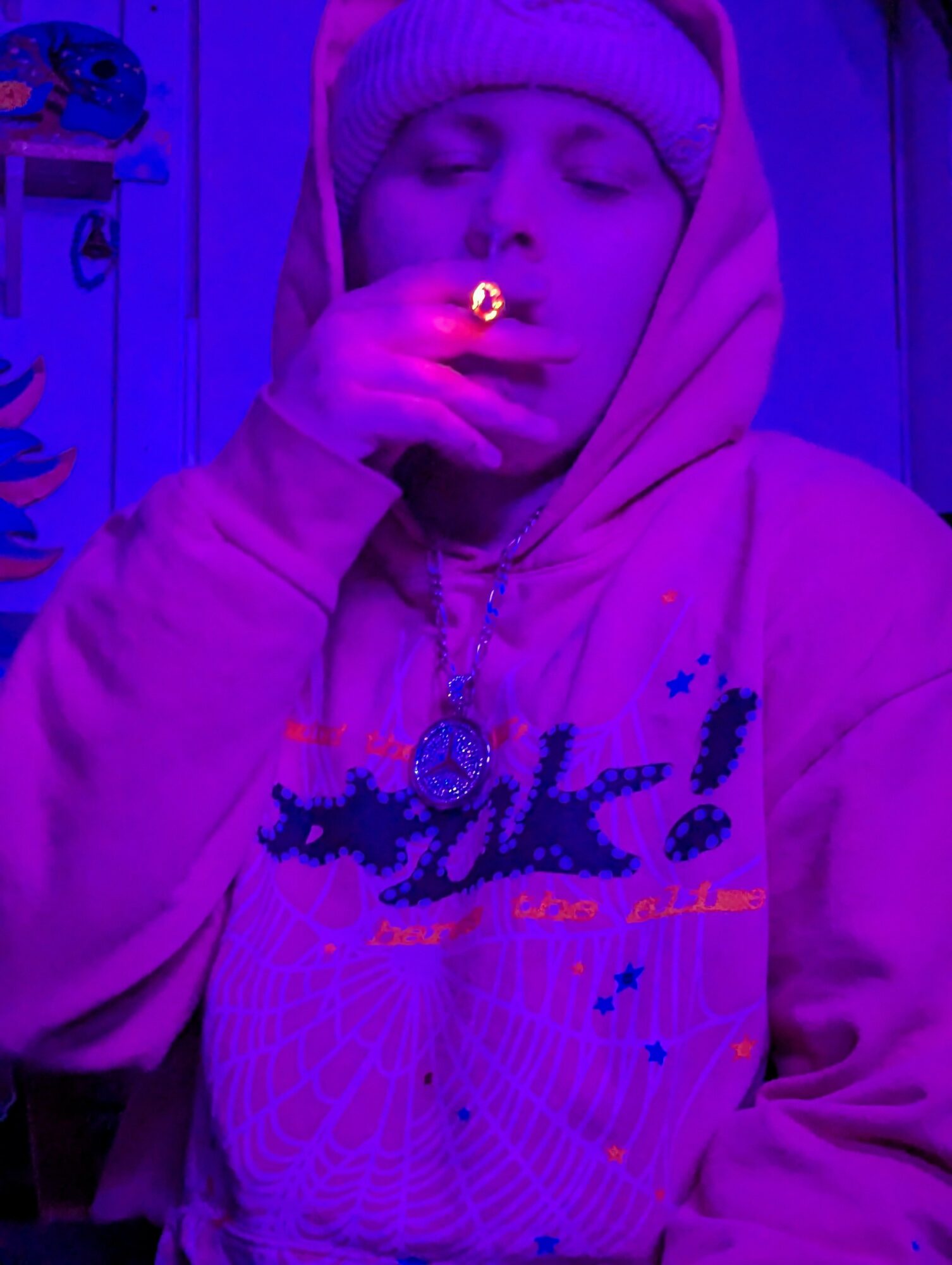 Person wearing hoodie and beanie, smoking, illuminated by purple and blue lighting, eyes closed, holding cigarette near face.