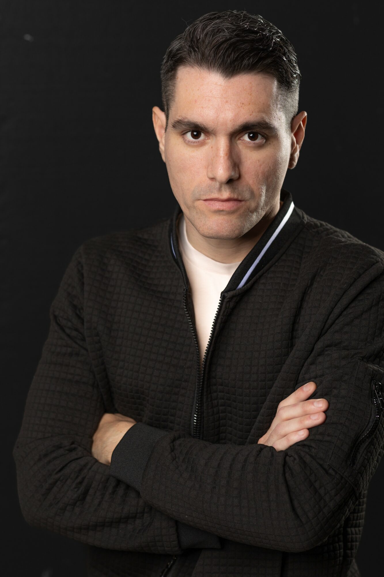 Man with dark hair and light skin, wearing a black jacket, crossing arms, against a black background.