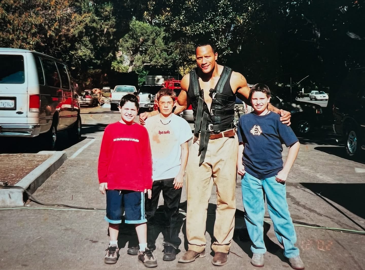 Man with three children standing outdoors in a parking lot, smiling, with trees and cars in background.