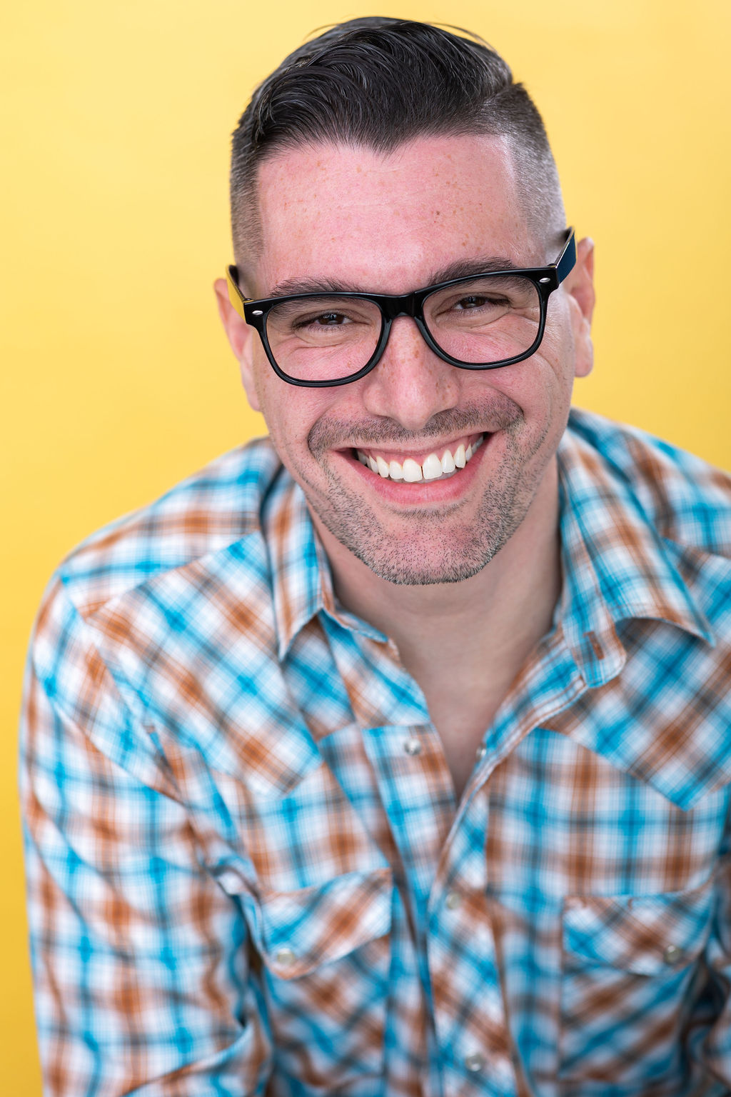 Smiling man with glasses and short dark hair, wearing a plaid shirt, against a yellow background.