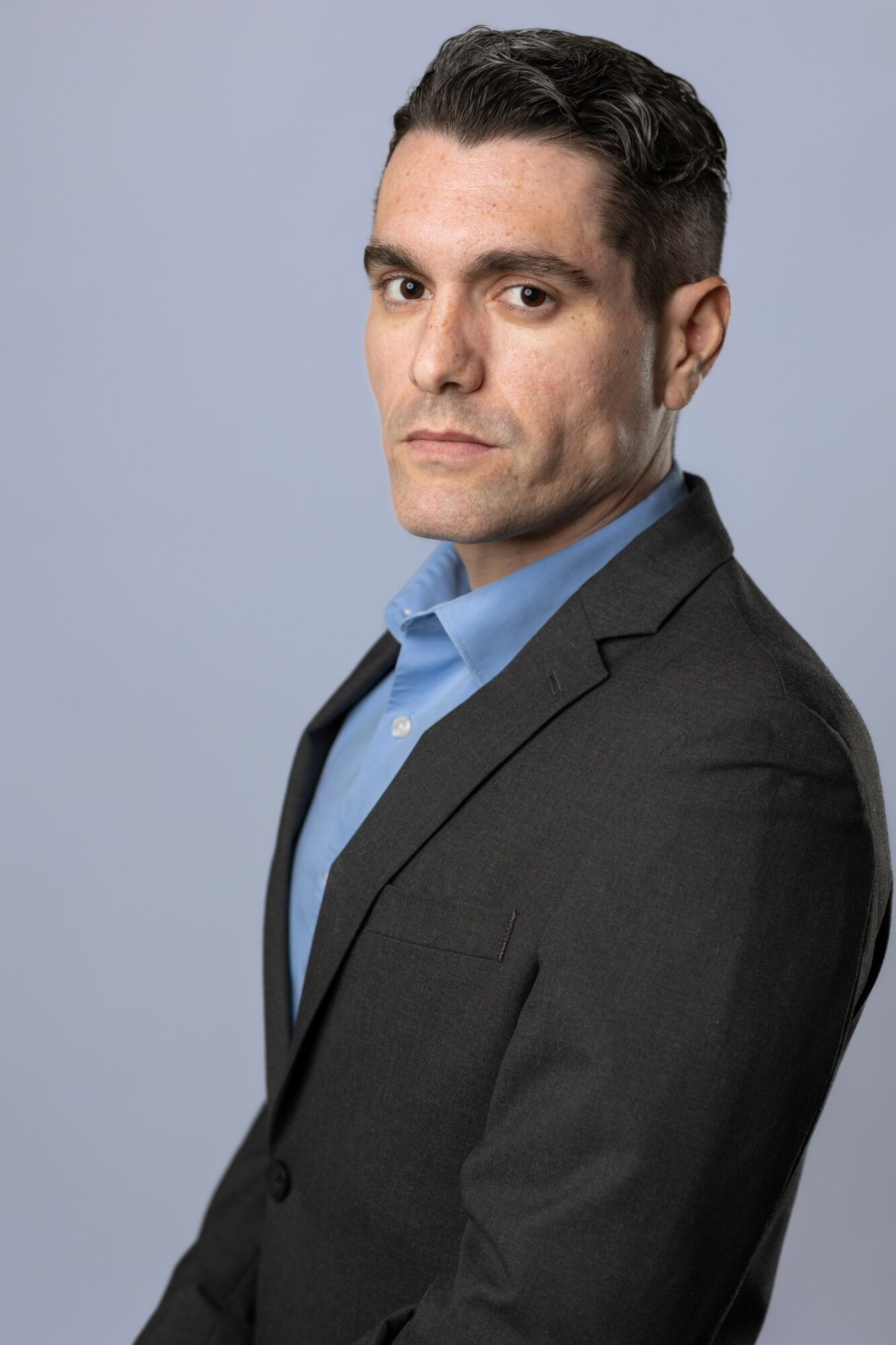 Man in a dark suit and blue shirt looking serious, facing slightly to the right, against a light gray background.