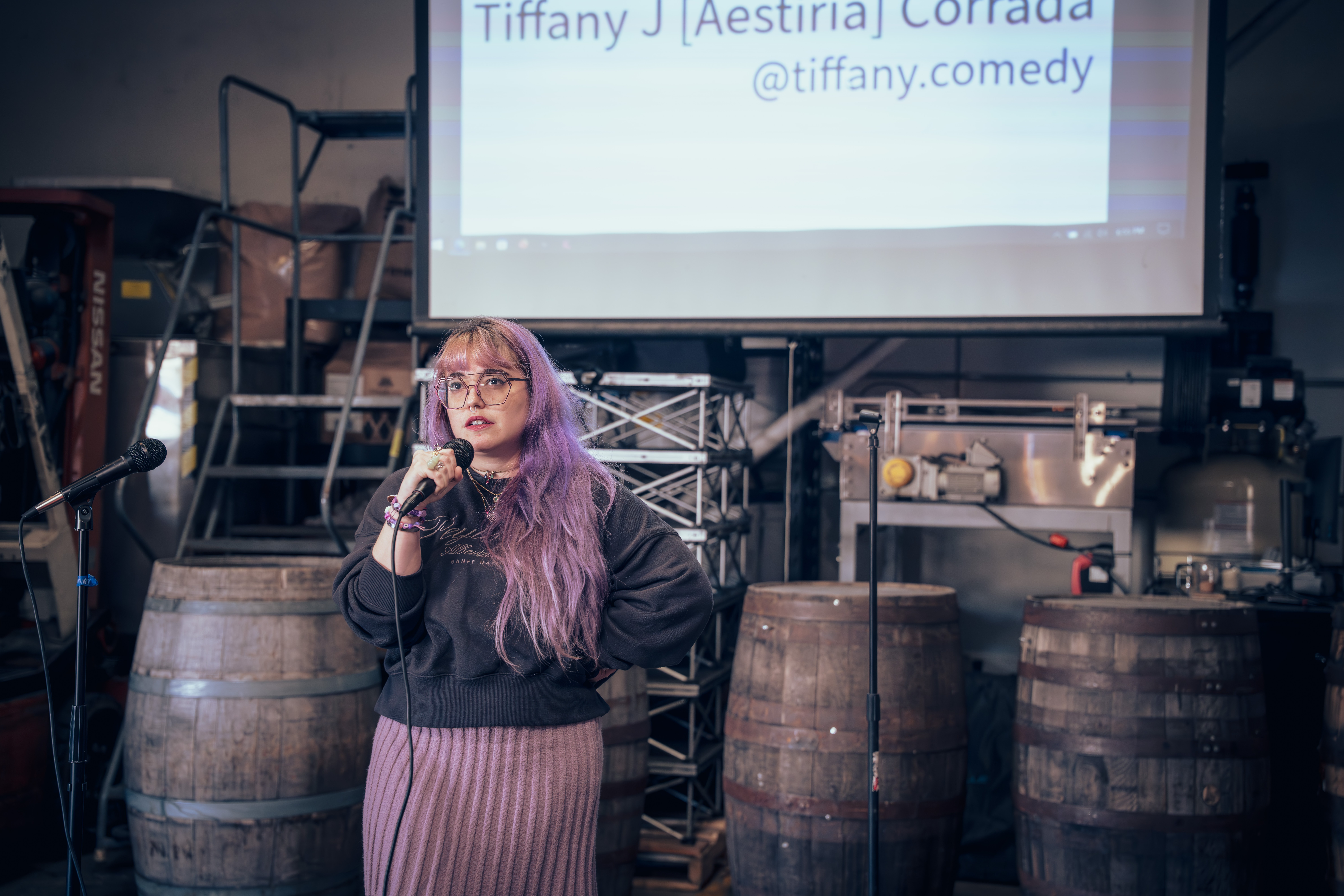 Woman with purple hair speaking into a microphone on stage, with barrels and a screen behind her.