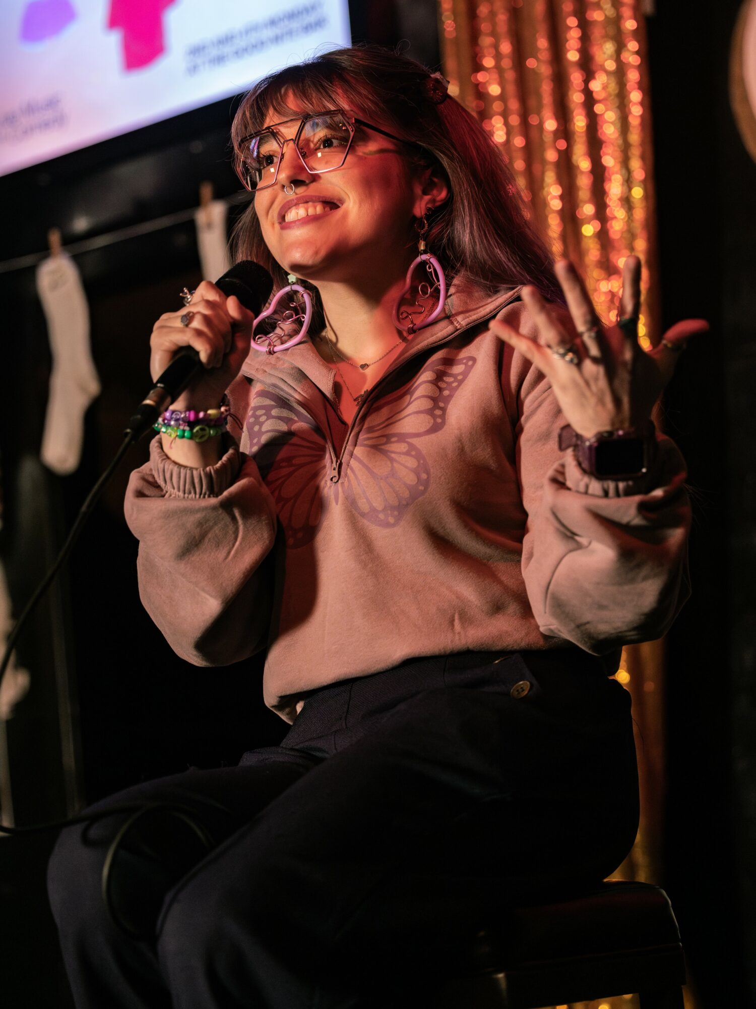 Woman with glasses smiling, holding a microphone, sitting on a chair, with a decorated background.