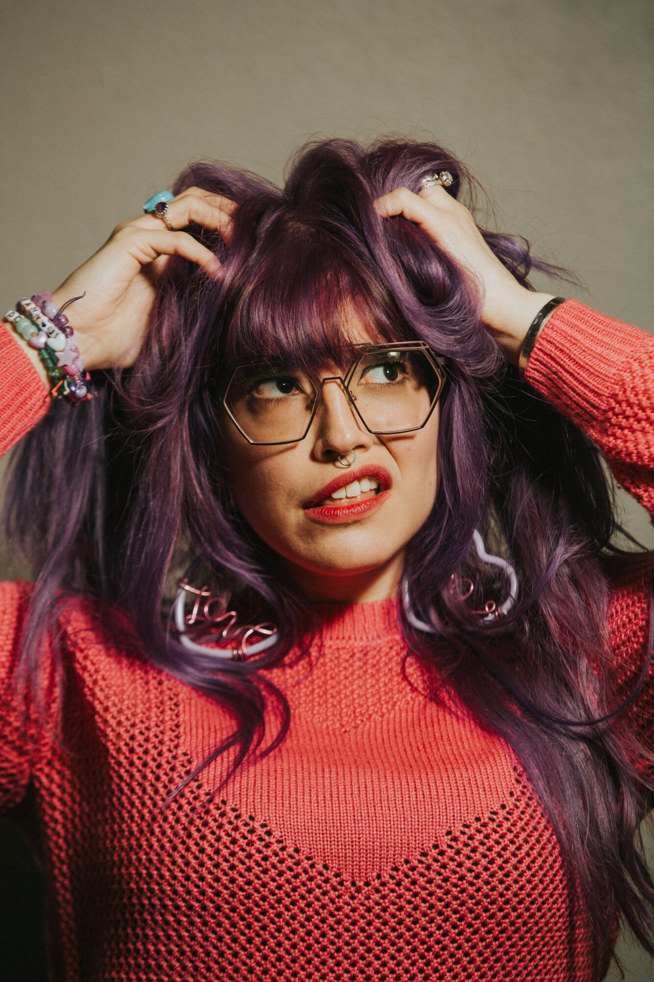Young woman with purple hair, glasses, and a red sweater, holding her head with a confused expression.
