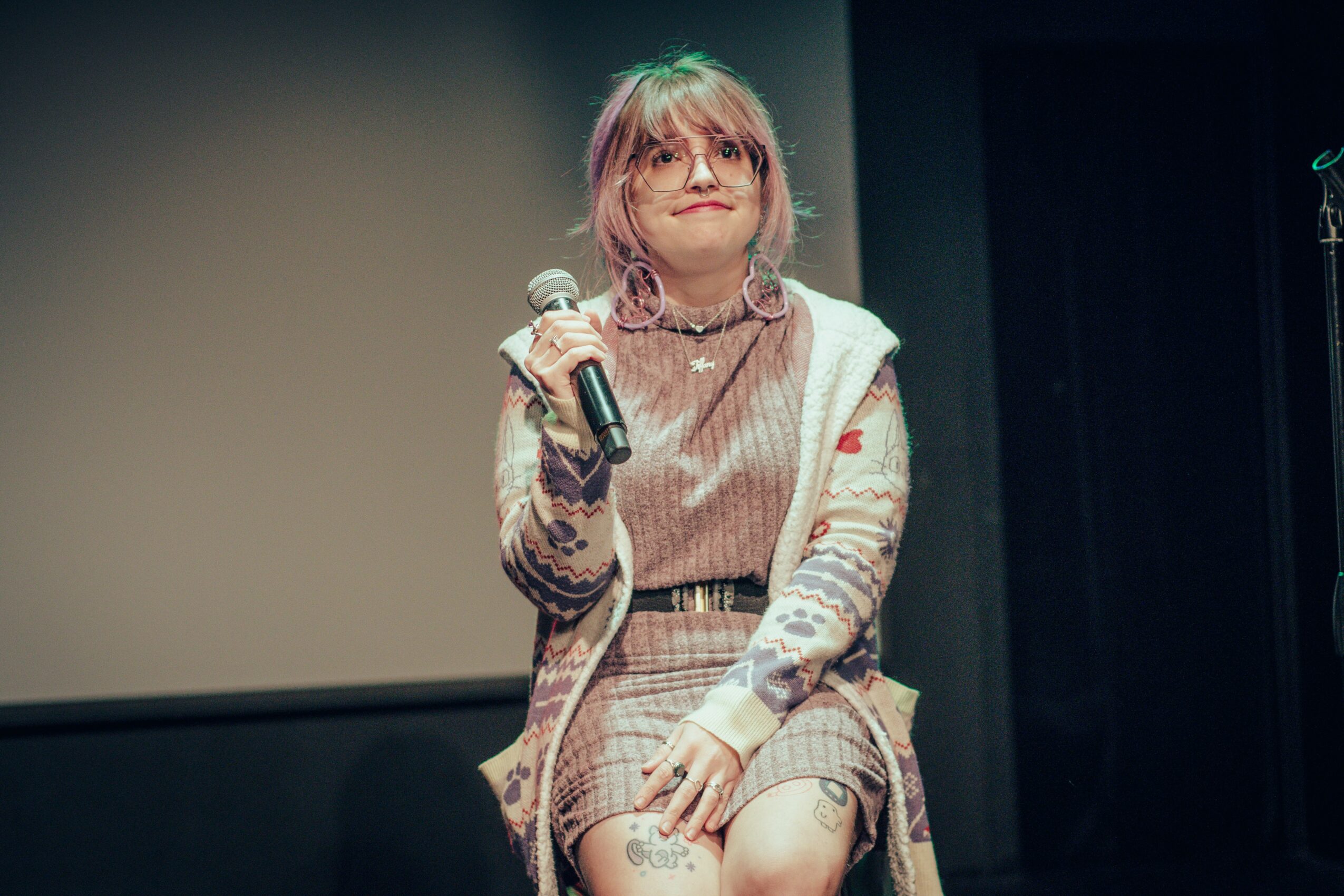 Person holding a microphone on stage, wearing glasses, a patterned sweater, and a plaid skirt.