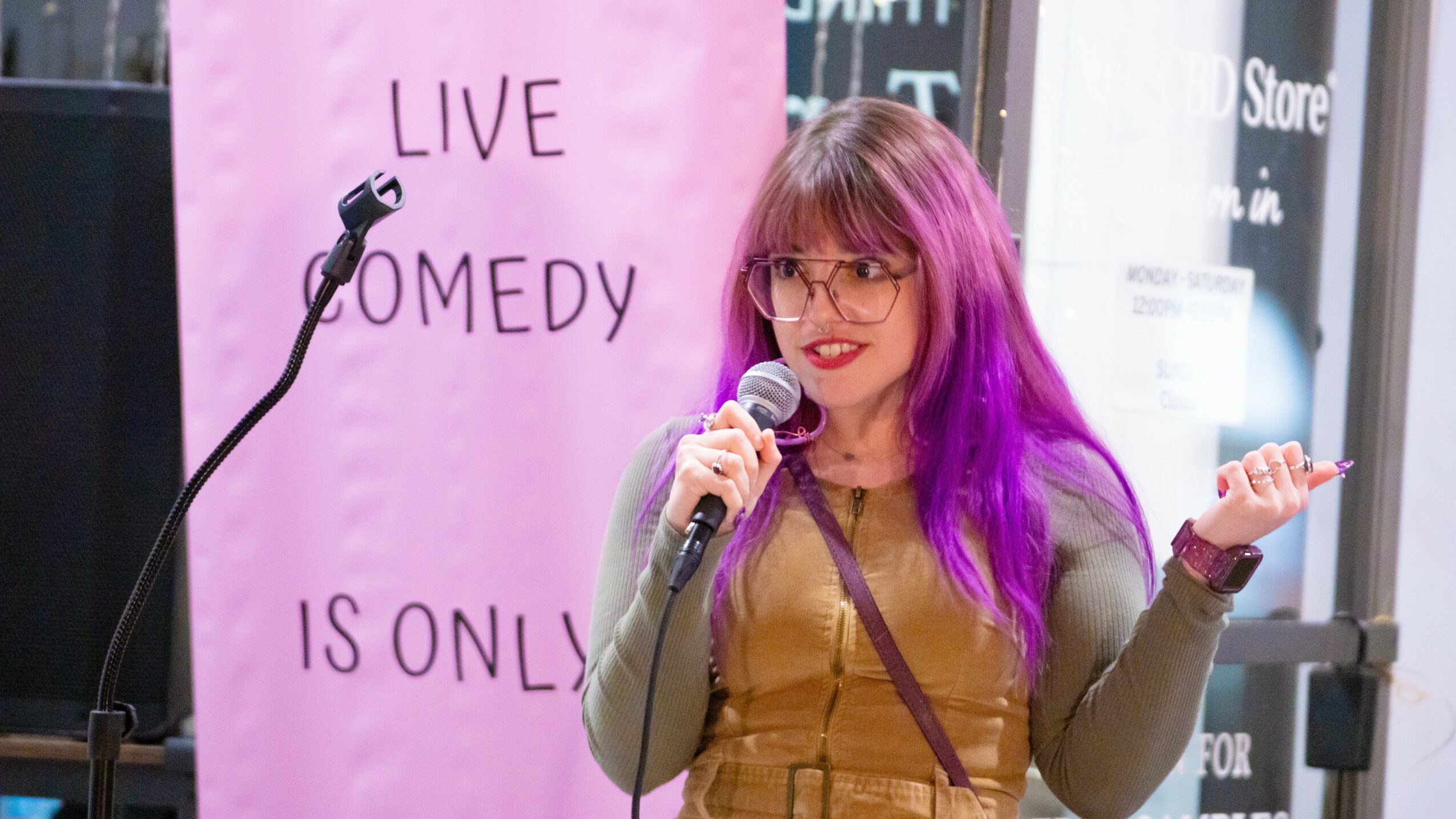 Woman with purple hair and glasses speaking into a microphone, standing in front of a pink banner with text.