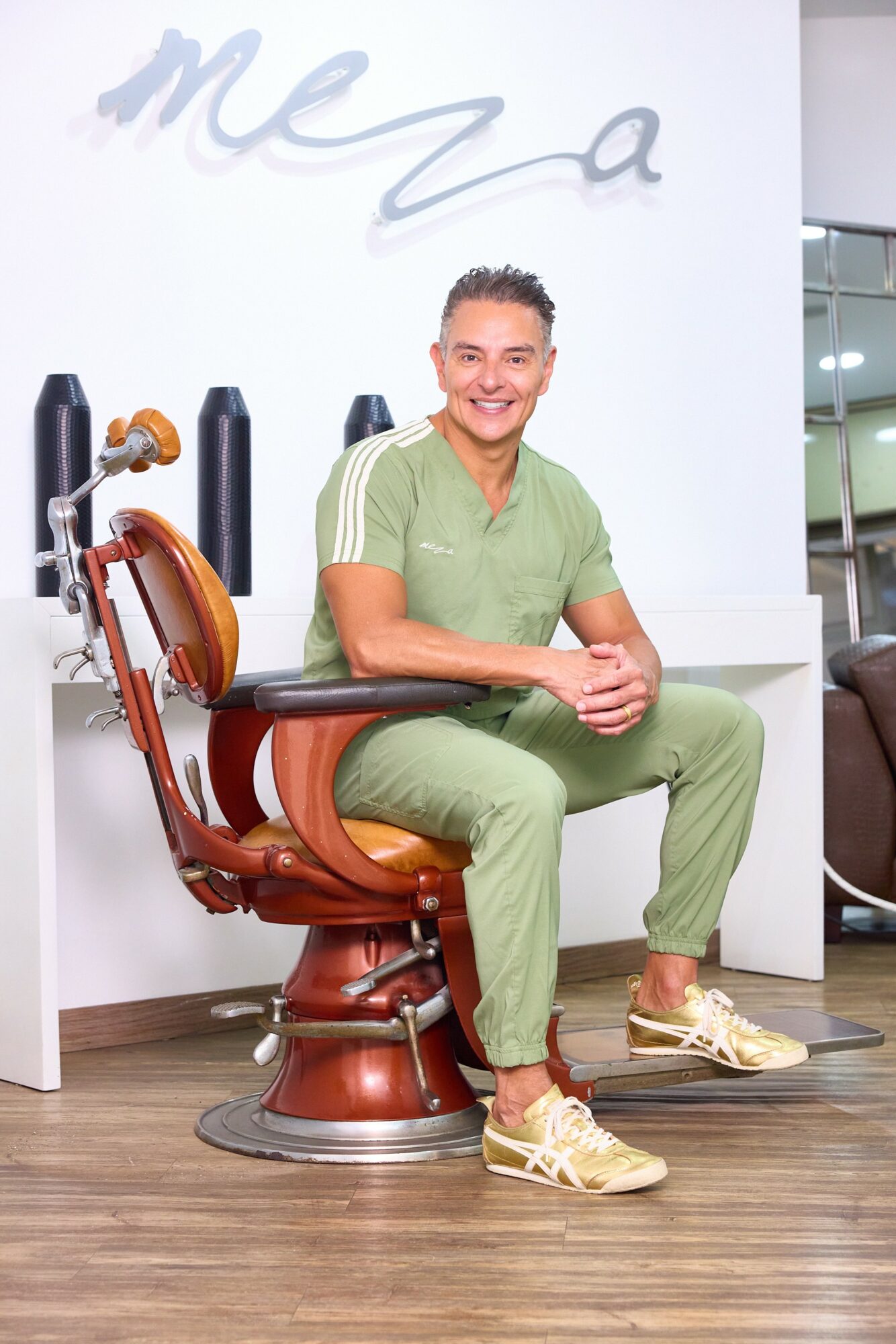 Person sitting on a vintage barber chair in a salon, smiling, wearing green scrubs and sneakers, with a white wall and salon equipment behind.