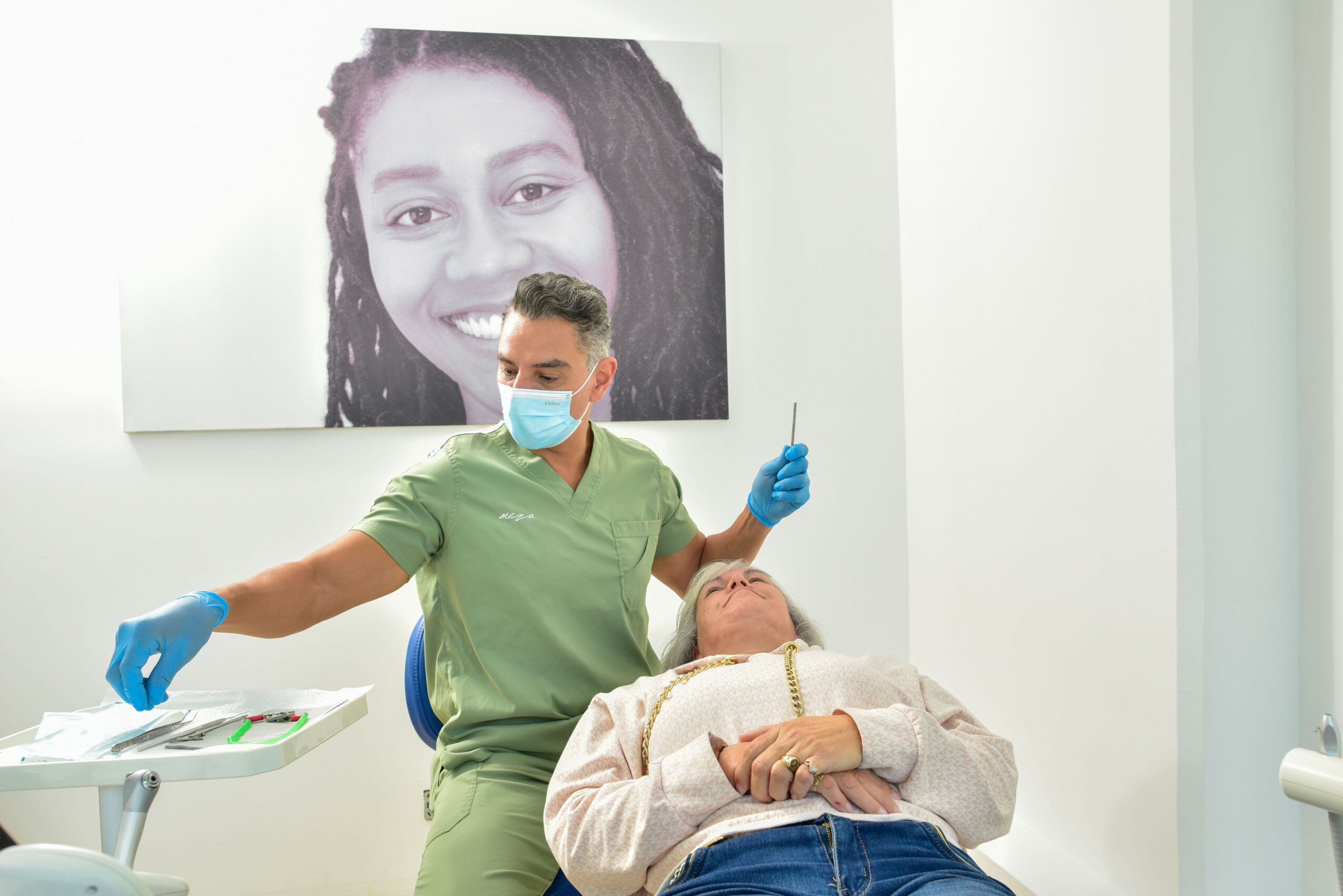 Dental professional in green scrubs and mask administers anesthesia to a patient lying in chair, large black-and-white portrait of a smiling woman in background.