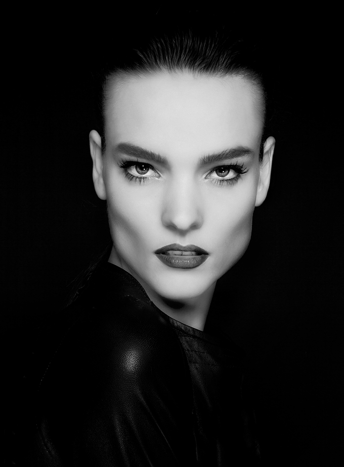 Black and white portrait of a woman with slicked-back hair, intense gaze, and dark lipstick, looking directly at the camera.