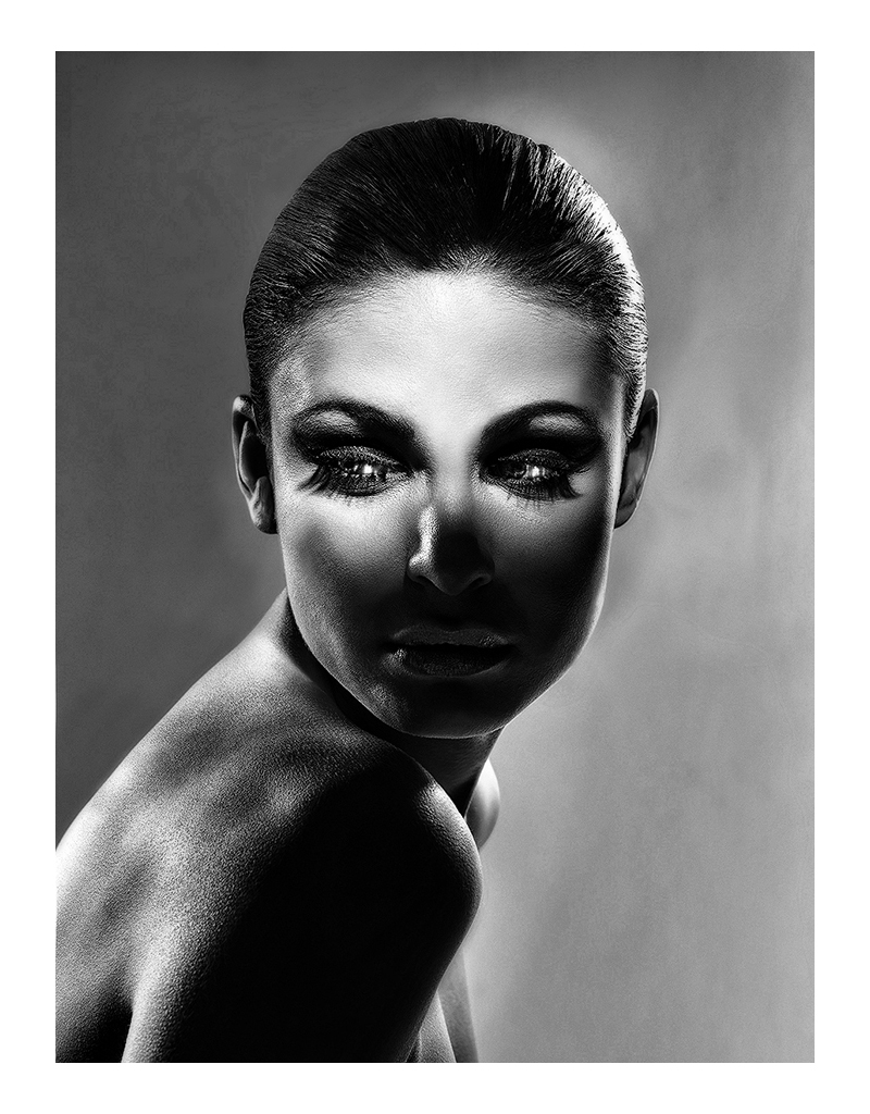 Black and white portrait of a woman with slicked-back hair, looking directly at the camera, with dramatic lighting highlighting her face and shoulder.