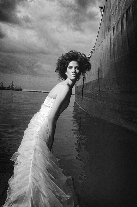 Woman in a flowing dress leaning against water near a large ship, cloudy sky in background.