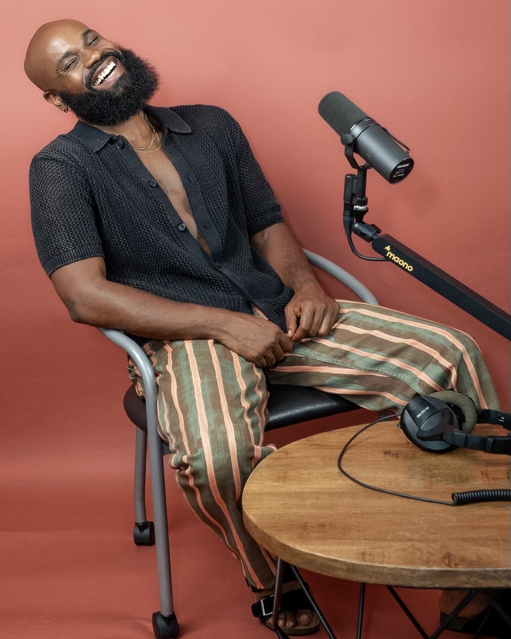 Man with a beard smiling, sitting in a chair with a microphone and headphones on a wooden table, pink background.