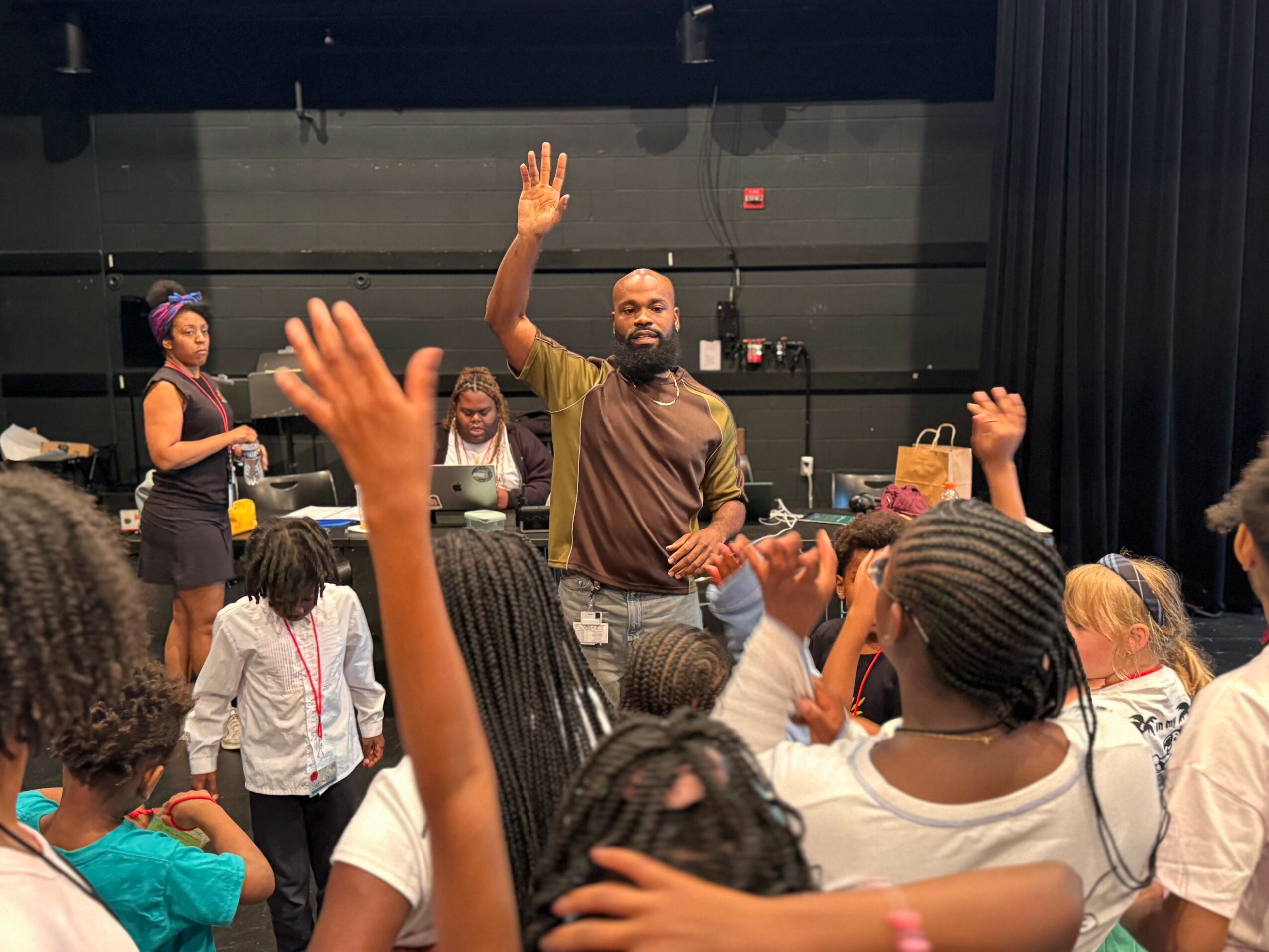Man with beard and brown shirt raising hand in front of children and adults in a room.