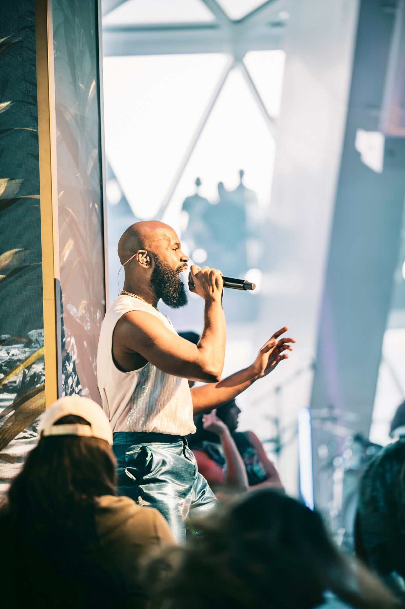Male performer with a beard singing into a microphone on stage, audience in foreground, modern architectural background.