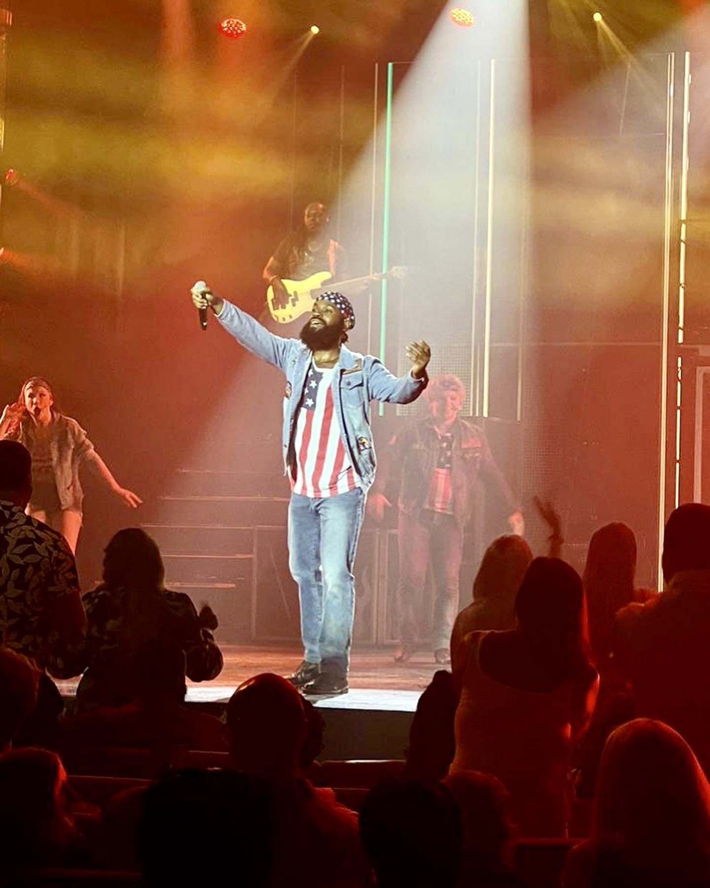 Performer on stage with arms raised, audience in foreground, colorful stage lights, musician playing guitar in background.