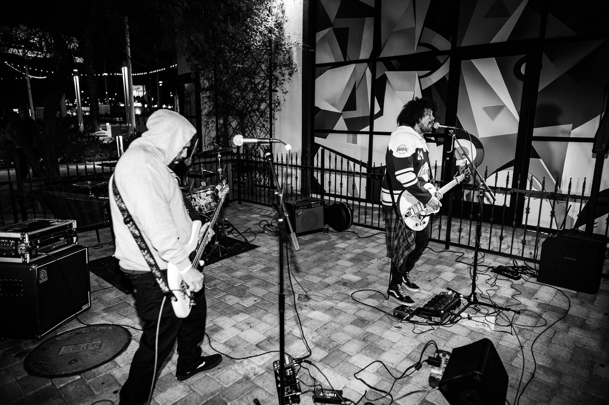 Two musicians perform with guitars on a stage, one wearing a hoodie, in front of a colorful geometric backdrop.
