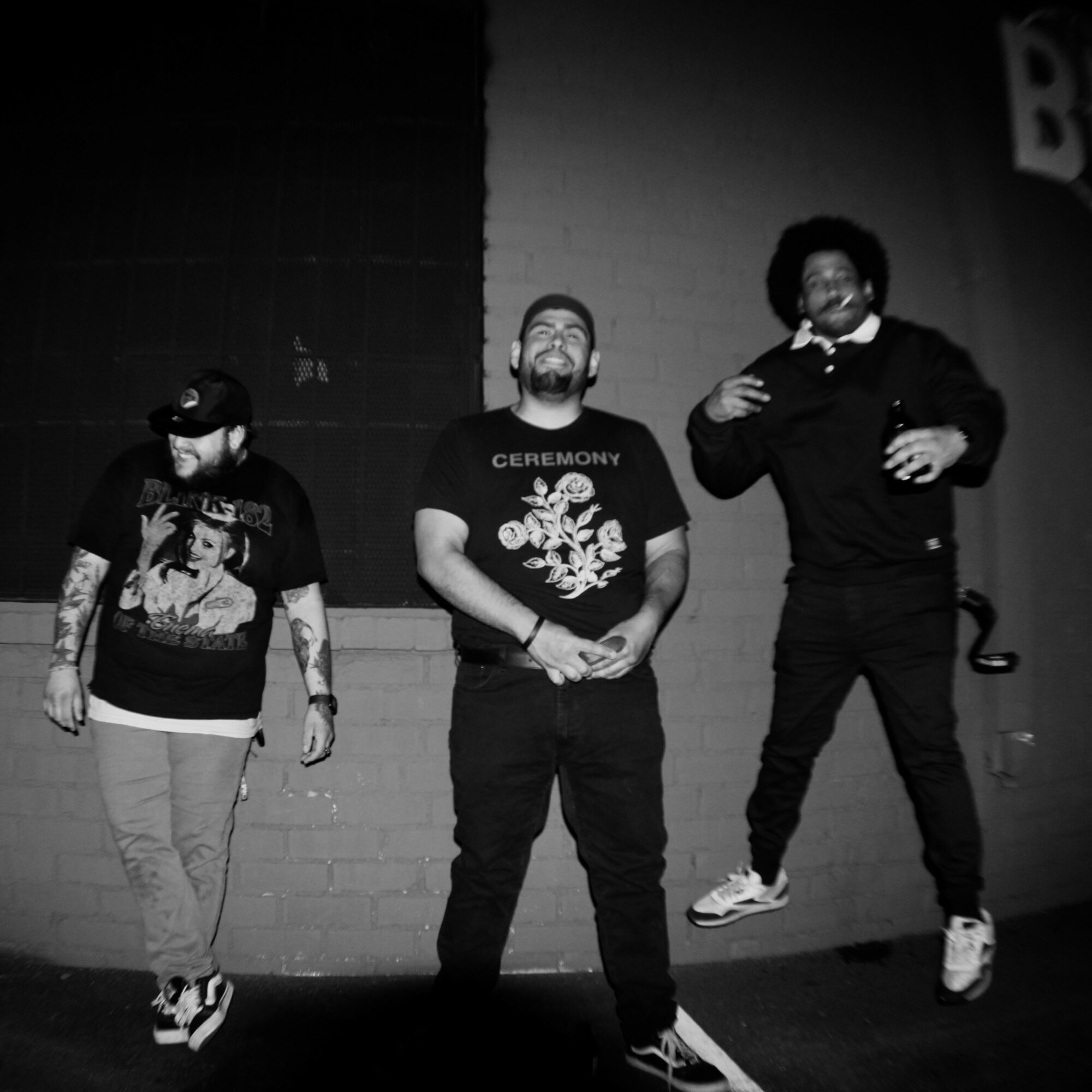 Three people standing on stage, two are smiling and one is looking down, with a brick wall background.