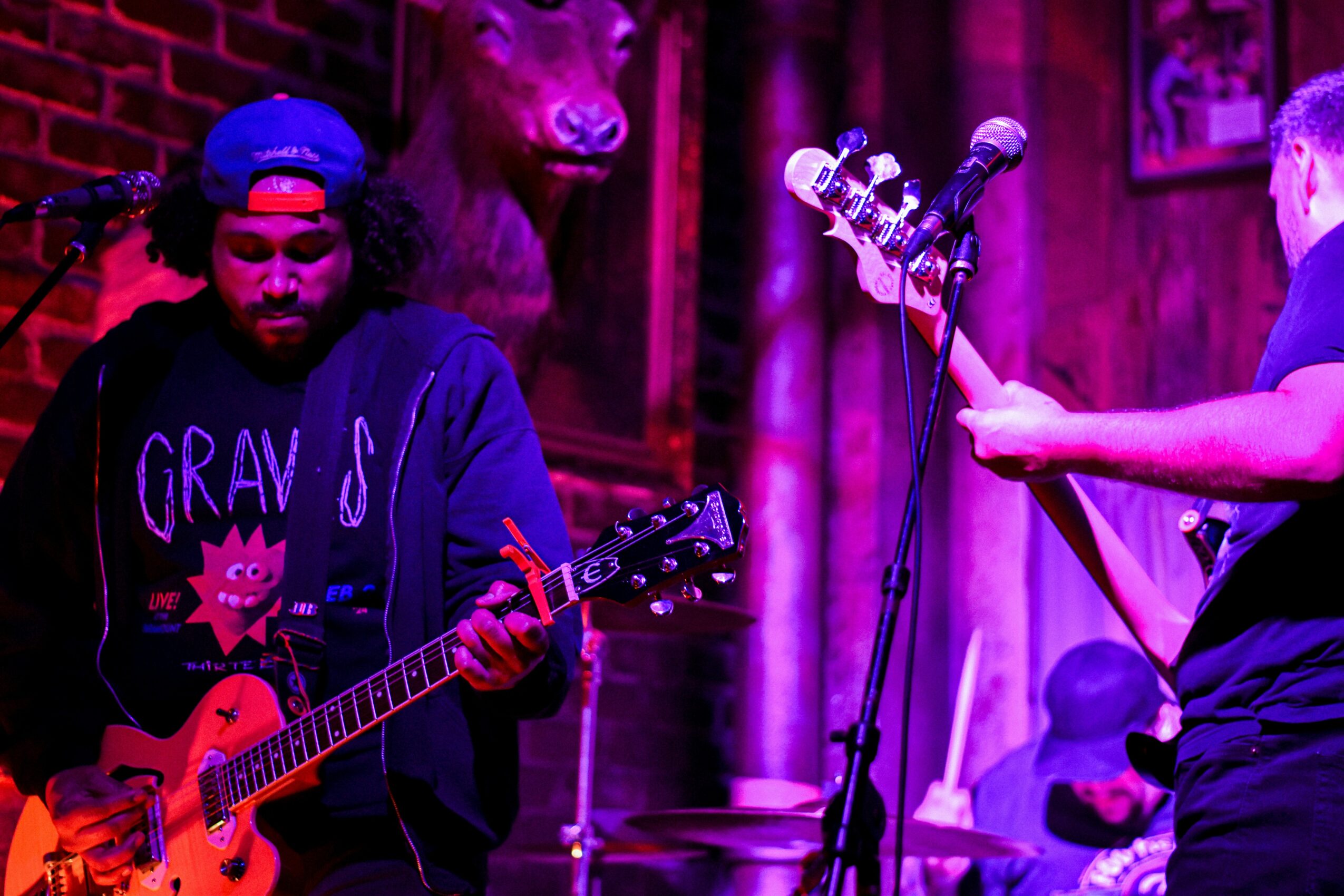 Two musicians perform on stage with guitars, one wearing a hoodie and the other with a cap, in a dimly lit venue.