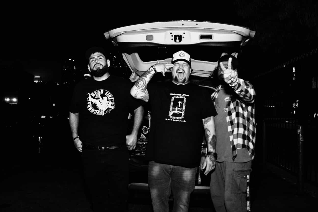 Three men standing outdoors at night, one in the center with a beard and tattoos, holding a skateboard above his head.