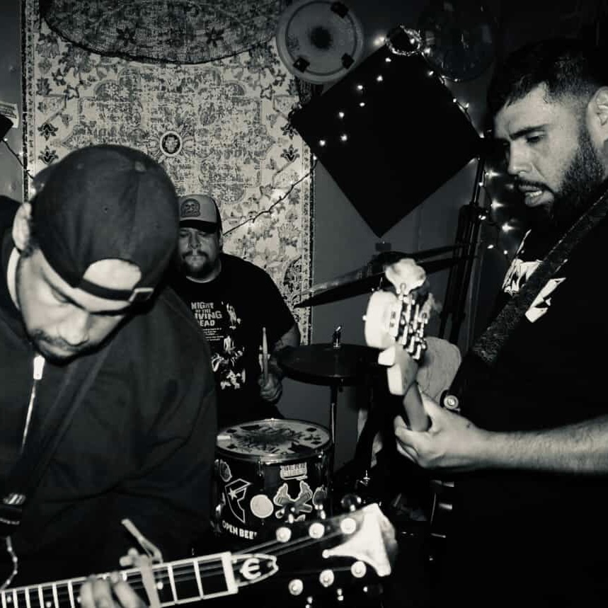 Three musicians playing guitars and drums in a dimly lit room with patterned wallpaper.