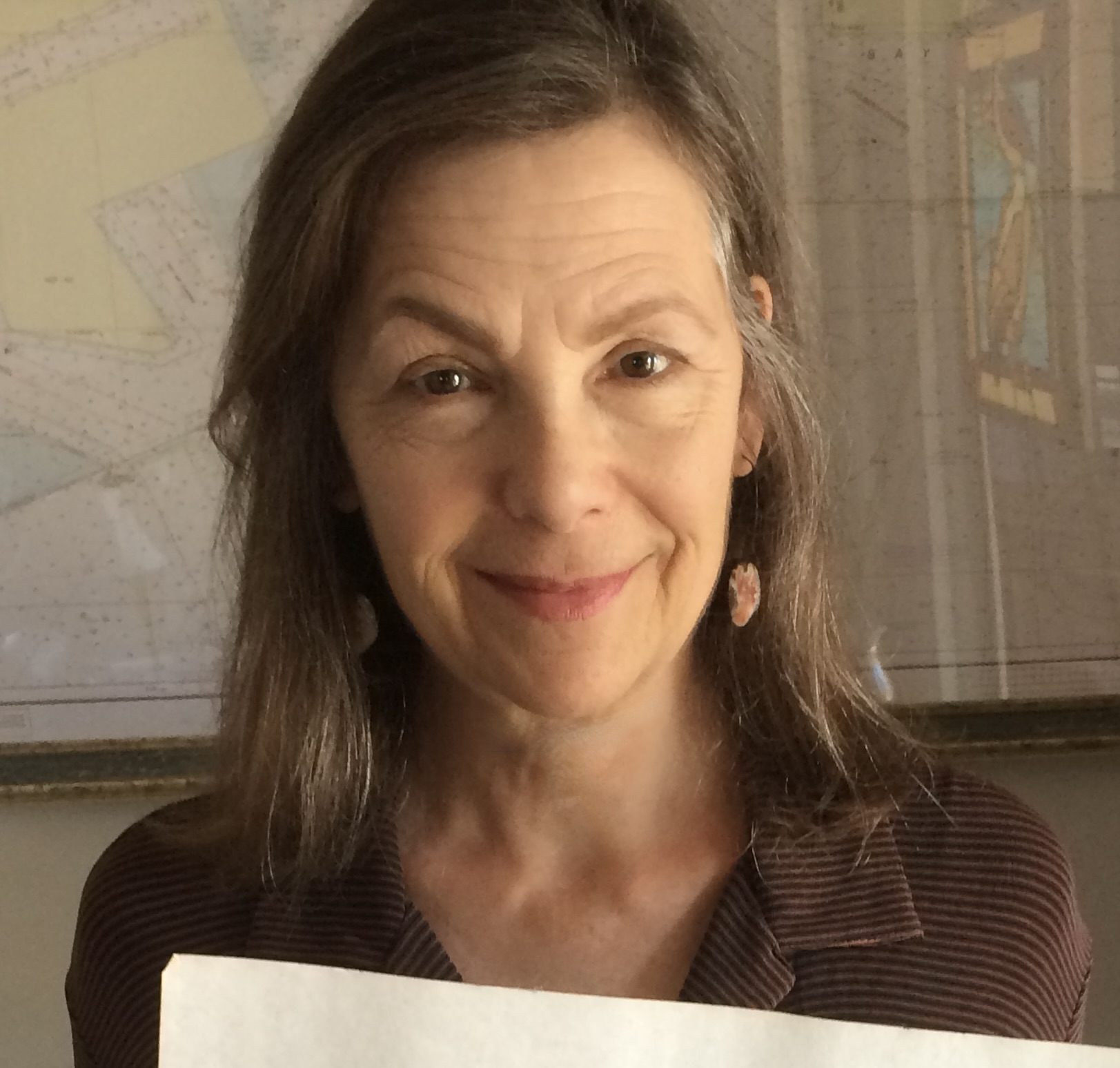 Woman with shoulder-length hair smiling, holding a paper, background with maps and charts