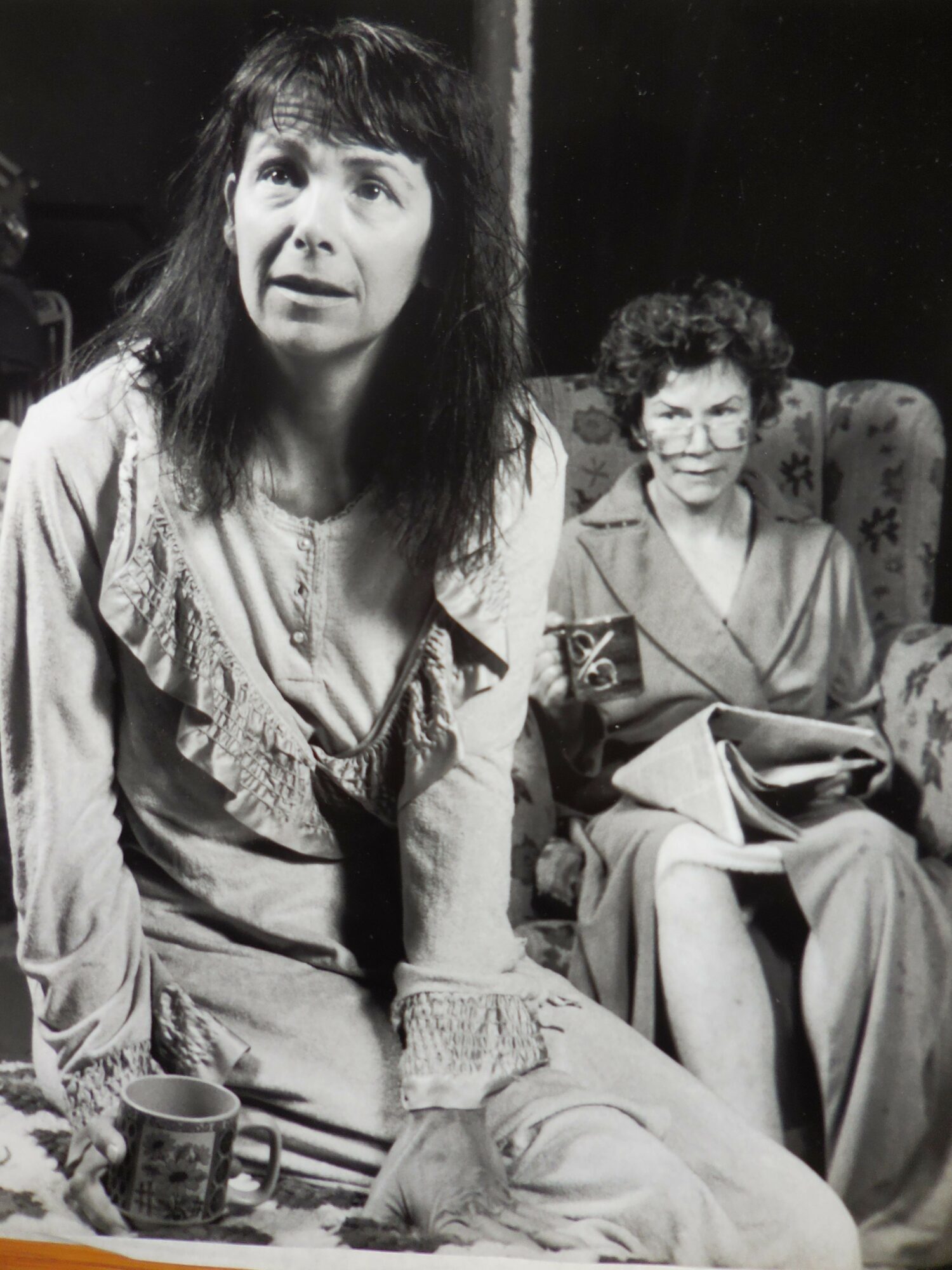 Two women sitting at a table, one with long dark hair and the other with curly hair, in a black and white photo.