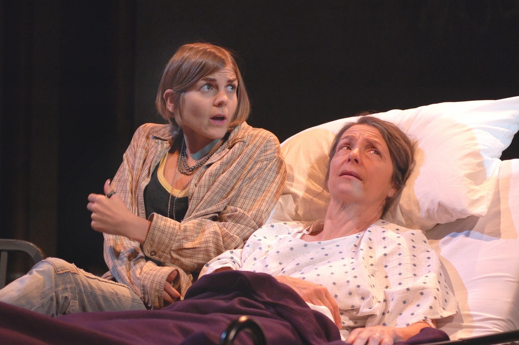 Two women on stage, one lying in bed and the other sitting beside her, both with surprised expressions.