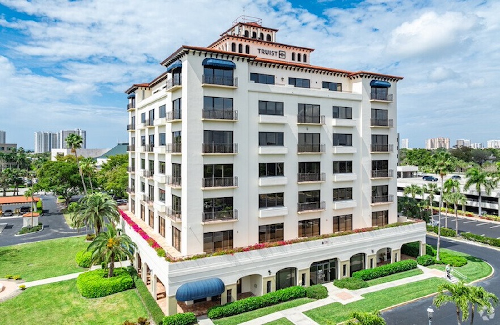 Multi-story building with balconies, surrounded by greenery and a parking lot, under a partly cloudy sky.