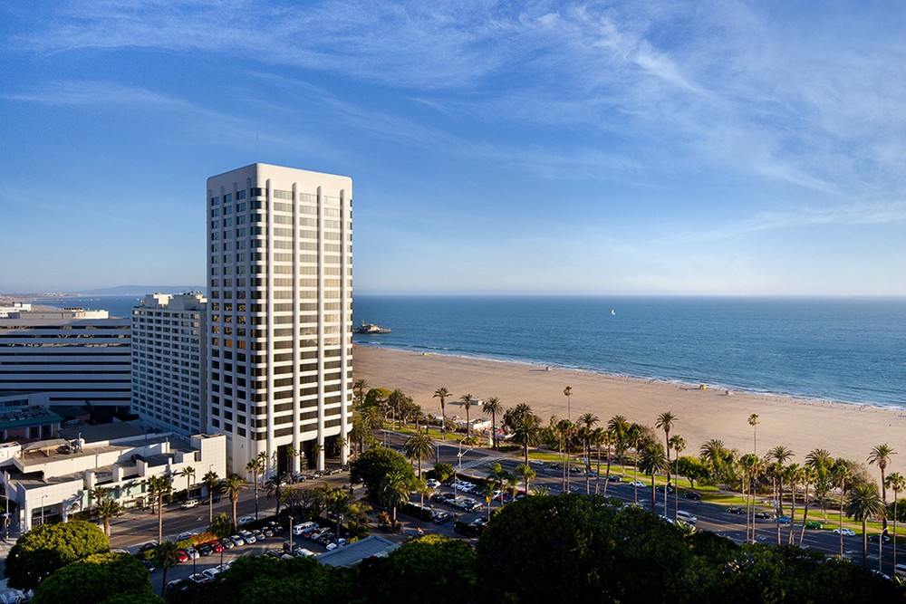 Tall white building near beach with palm trees, parking lot, and ocean in background under blue sky.