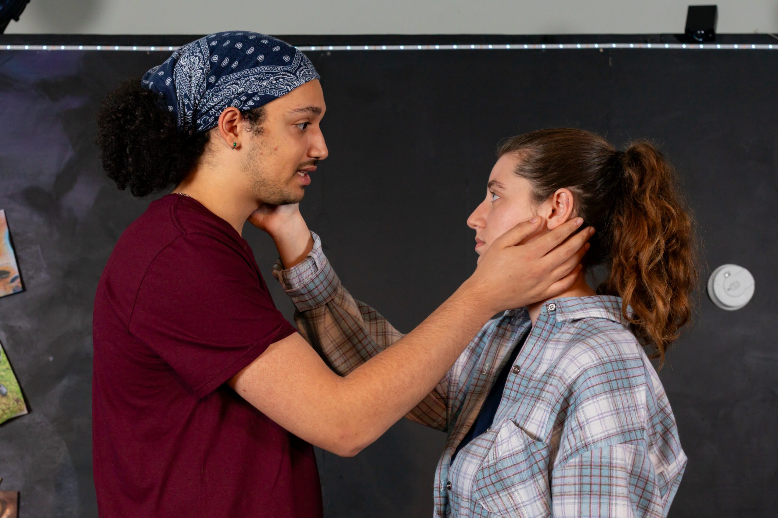 Two women face each other, one gently holding the other's face, in front of a blackboard.