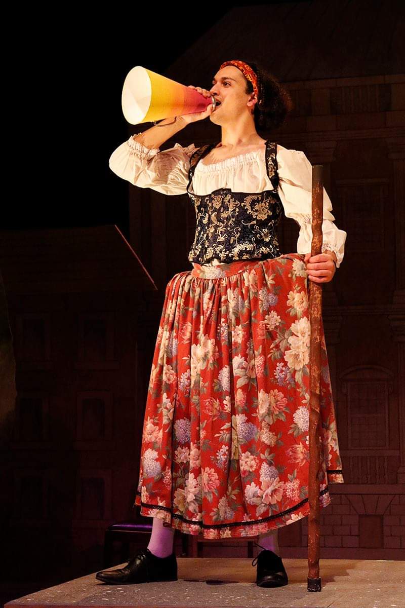 Woman in historical costume using a megaphone on stage, holding a staff, with a dark background.