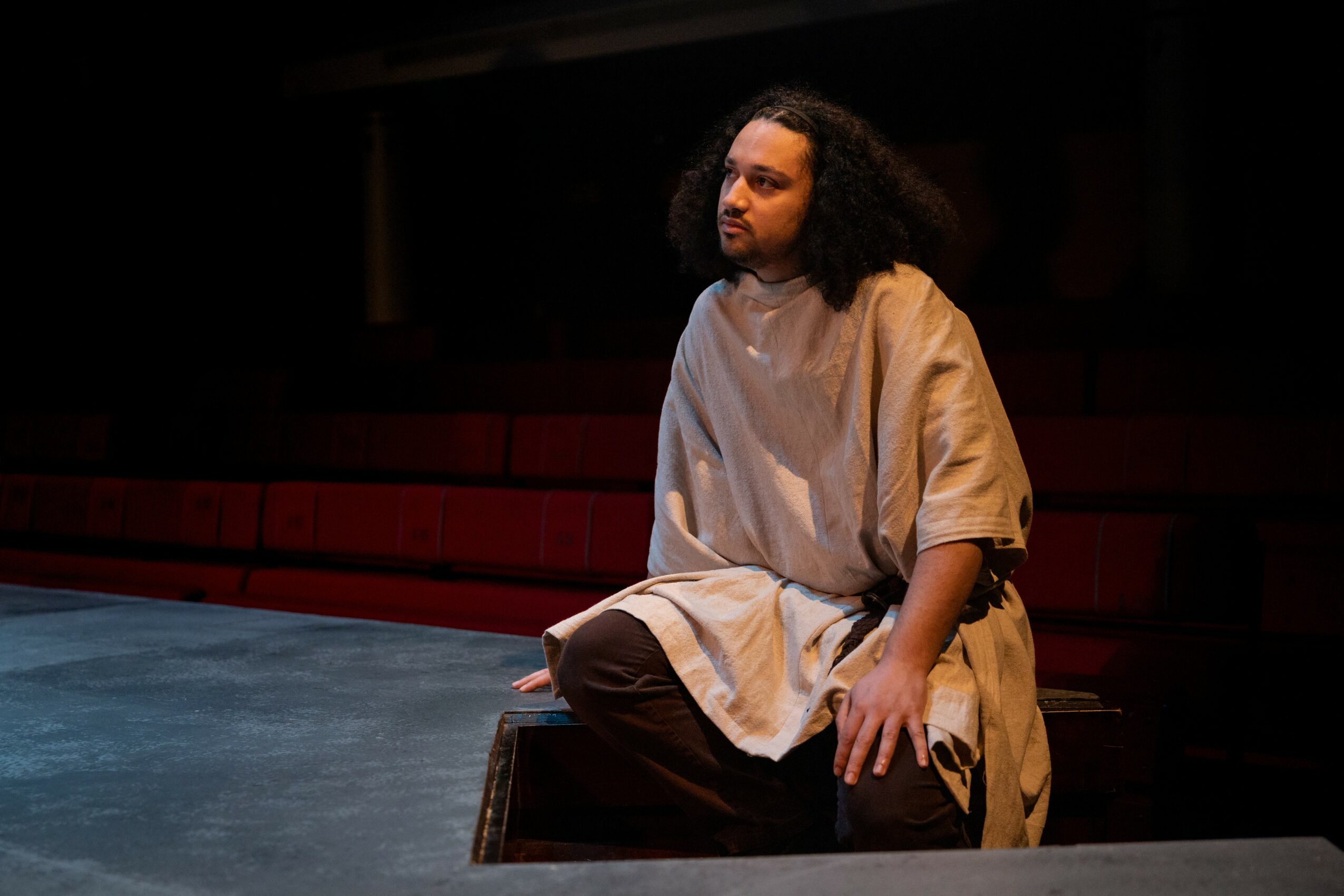 Person with curly hair sitting on stage in a beige costume, looking to the side, with a dark background and red seats.