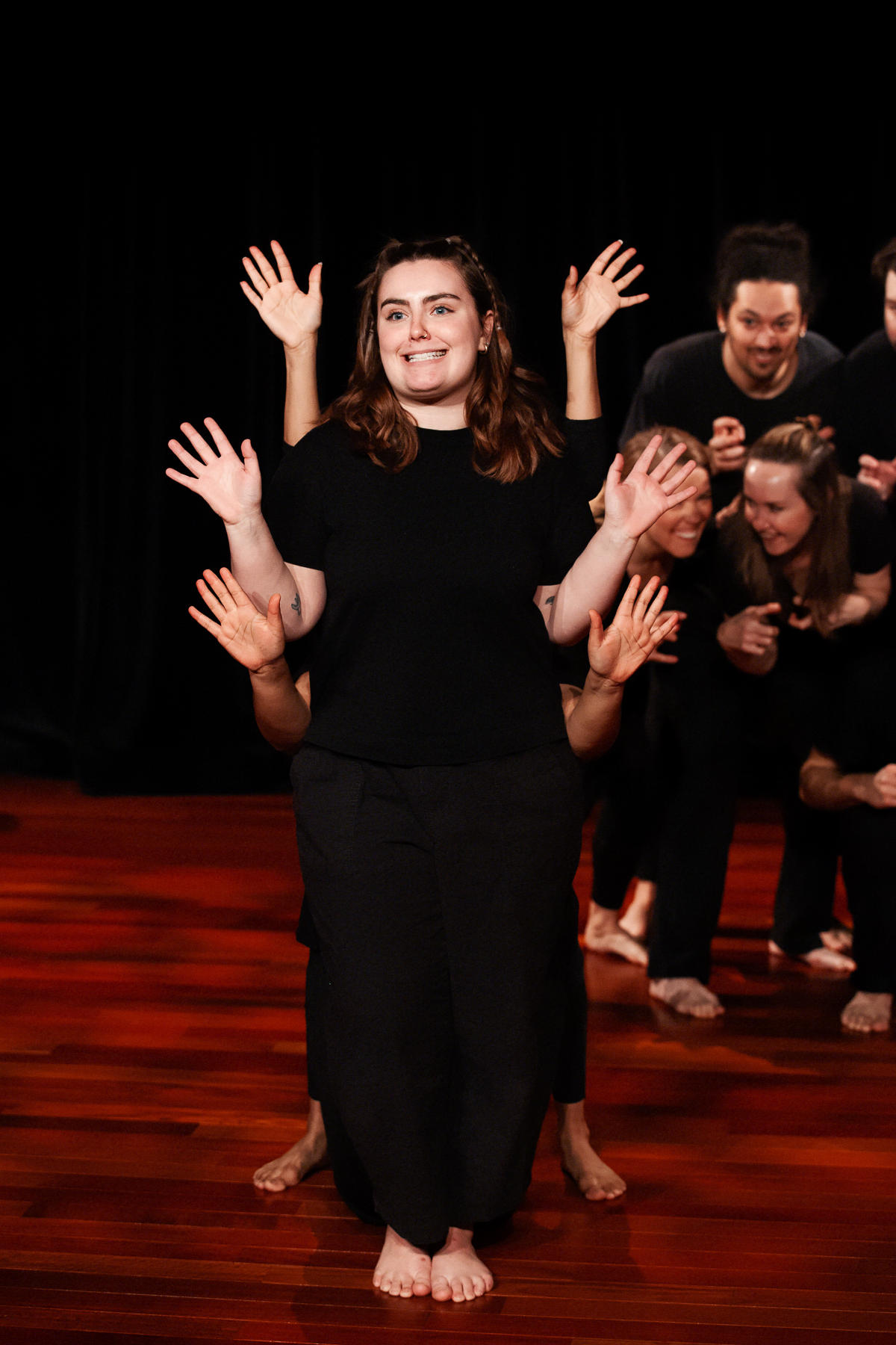 Group of people performing a dance or theatrical act on a wooden stage, with a woman in front smiling and raising her hands.
