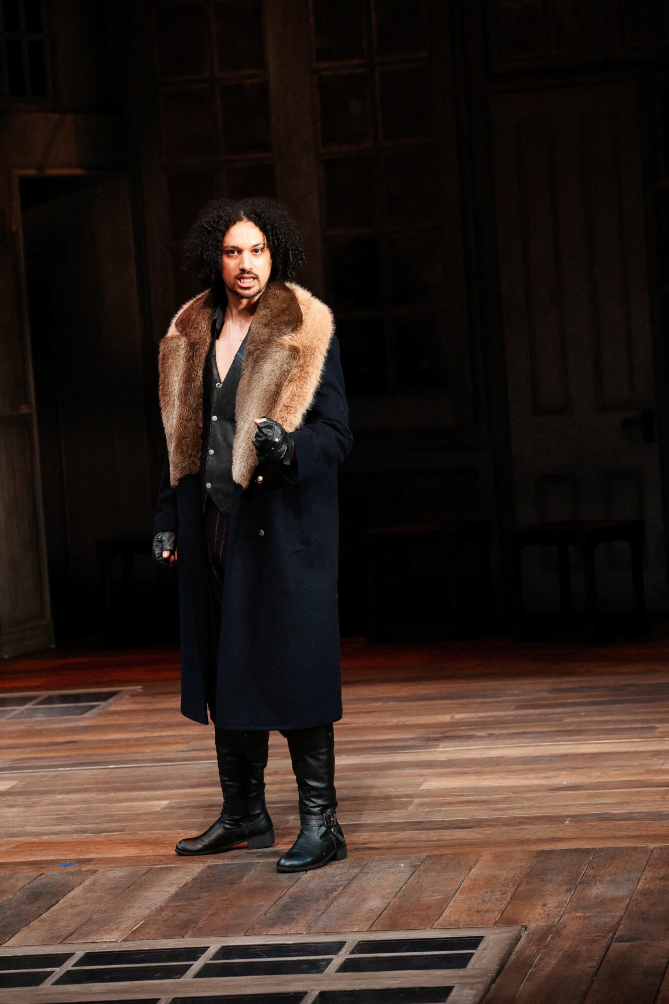 Person standing on wooden stage with dark background, wearing a long coat with fur collar, gloves, and boots.