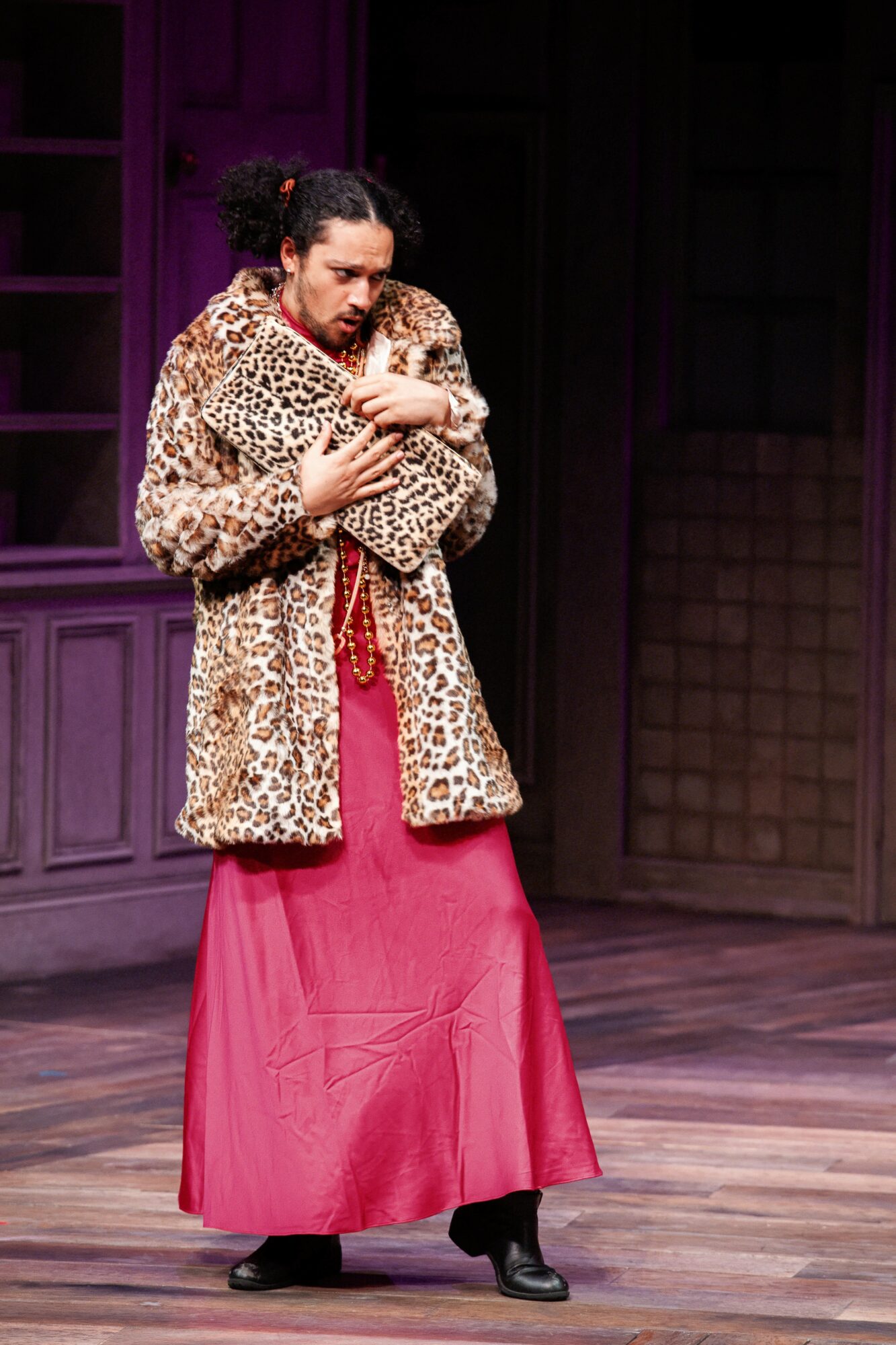 Person hugging a small dog, wearing a leopard print coat and a long pink skirt, on stage with wooden floor and dark background.