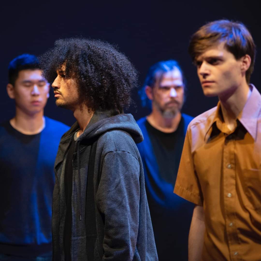 Four people standing in a dark room, some looking serious, with a woman with curly hair in the foreground.
