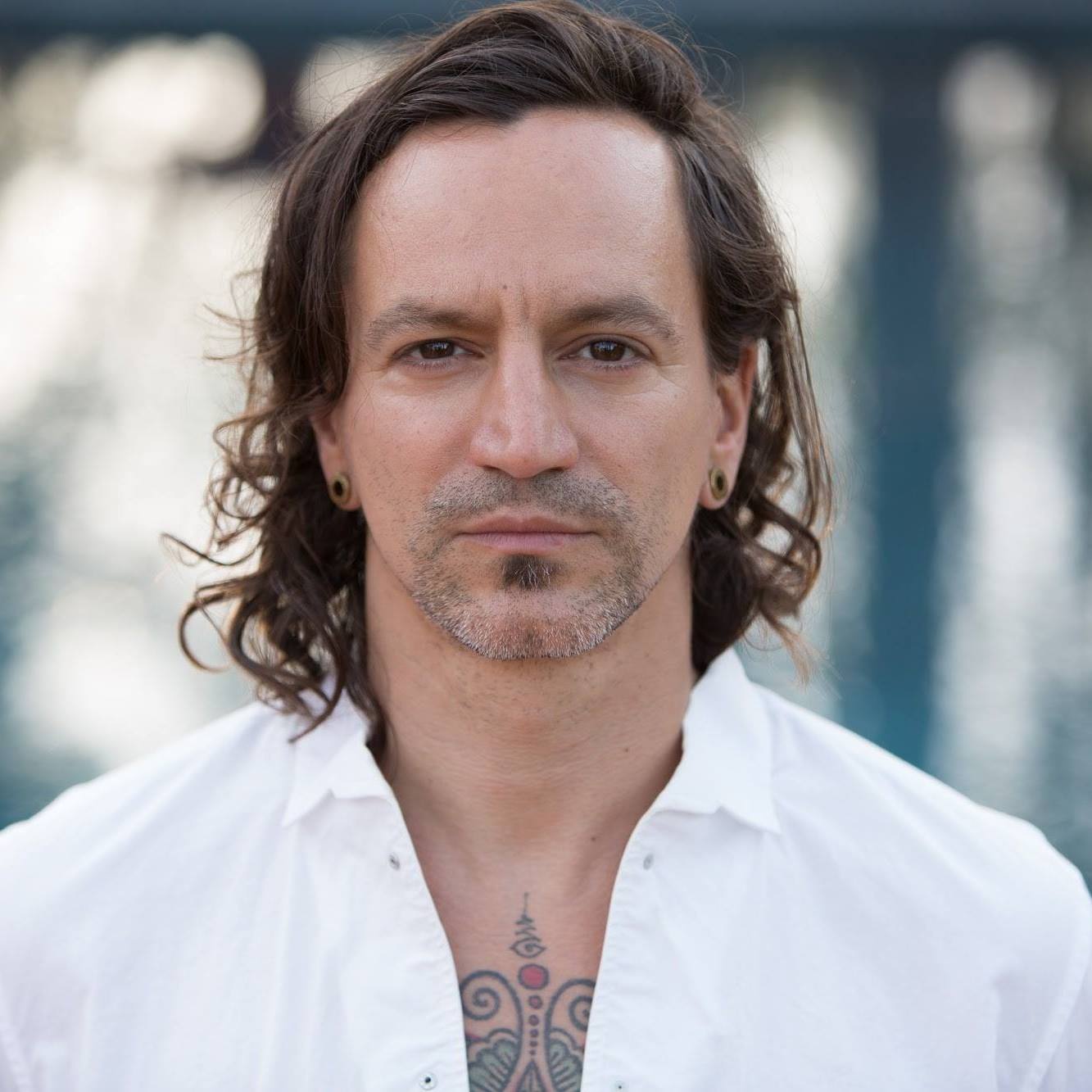 Man with long dark hair, light beard, wearing a white shirt, standing outdoors near water, with a blurred background.