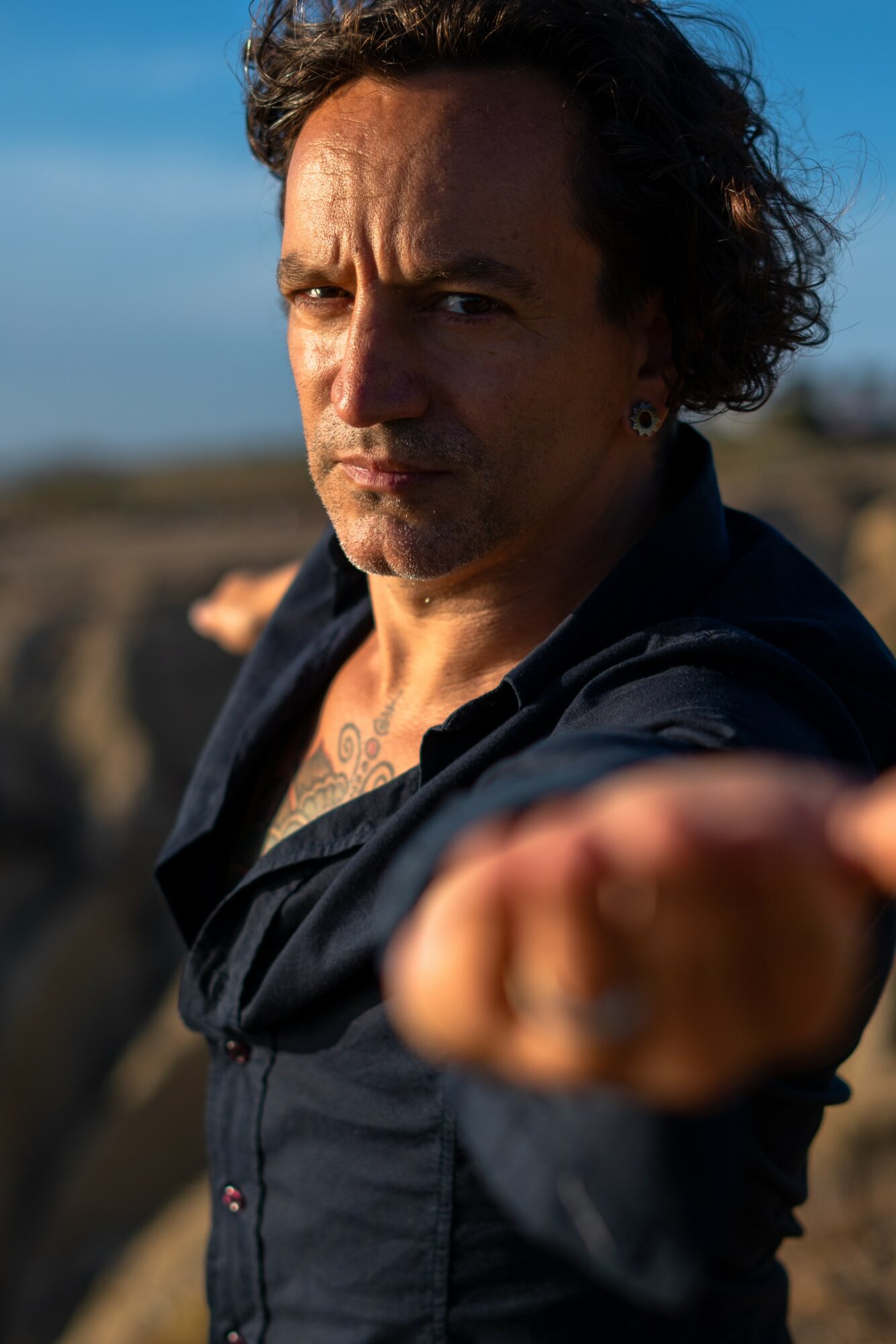 Man with curly hair and a dark shirt extends his arm toward the camera outdoors, with a blurred landscape background.