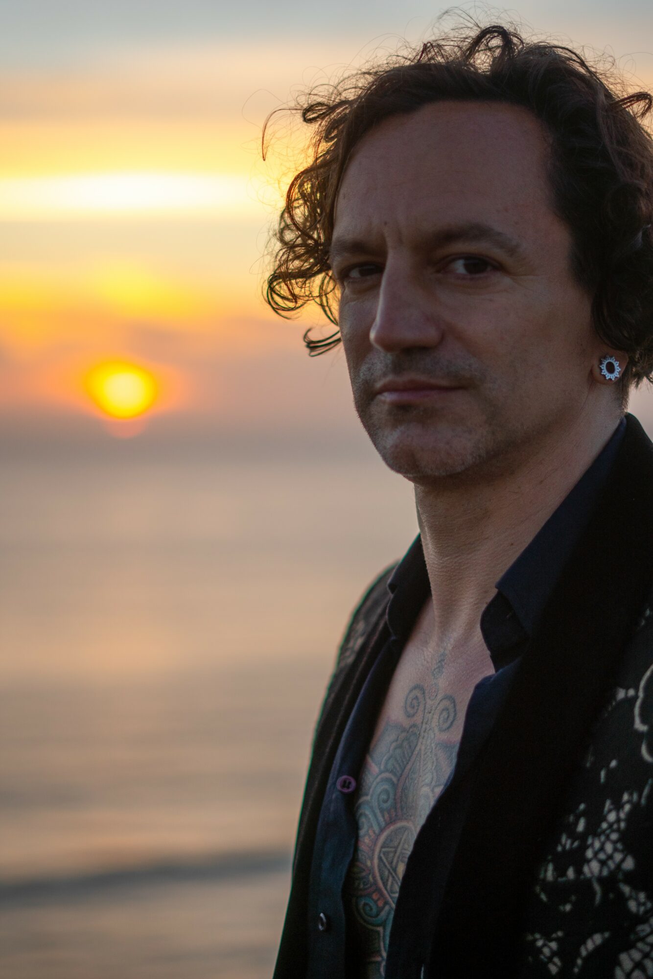 Man with curly hair and earrings stands outdoors at sunset, with a blurred ocean background.