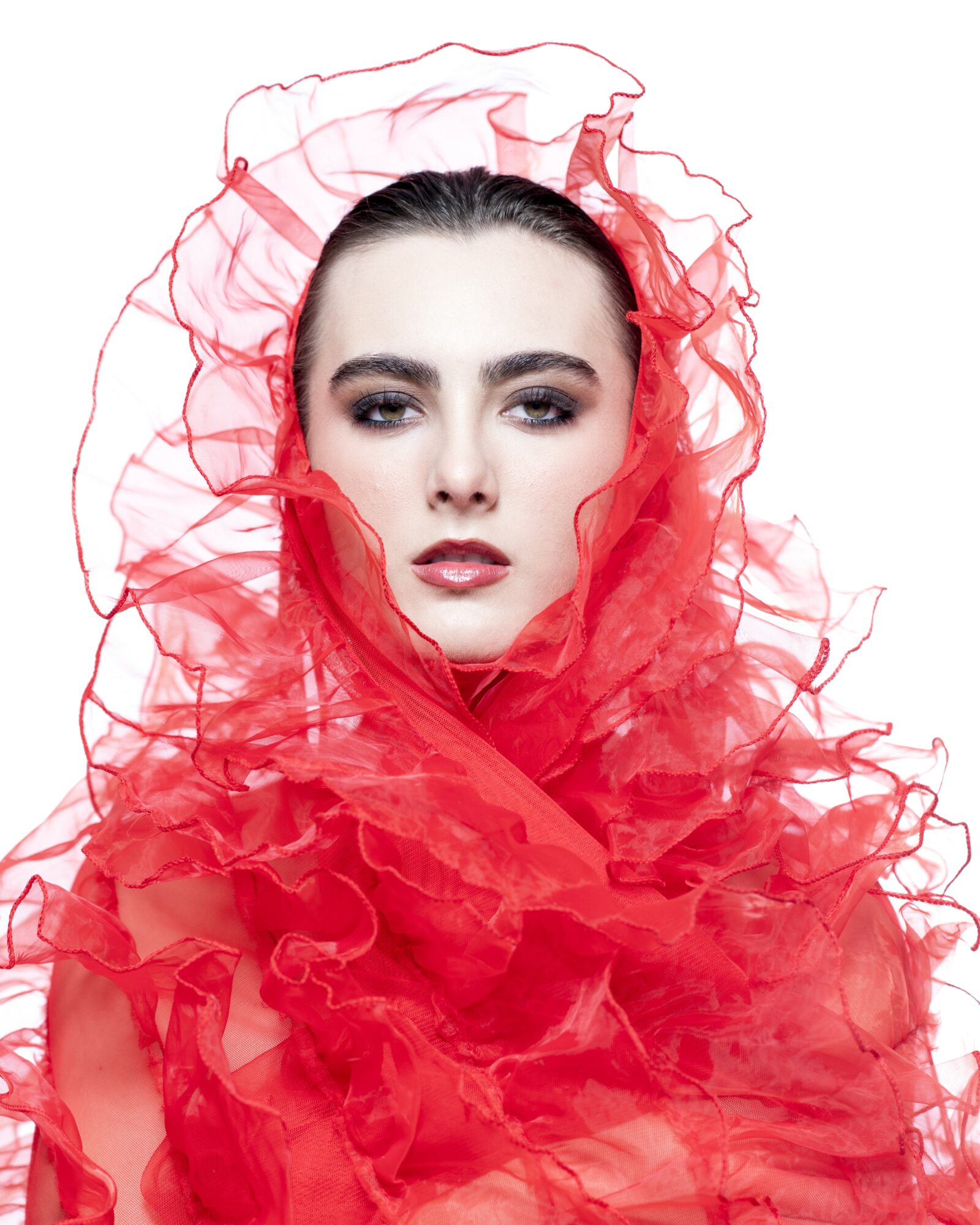 Woman with dark hair and makeup, wrapped in red ruffled fabric, facing forward against a white background.