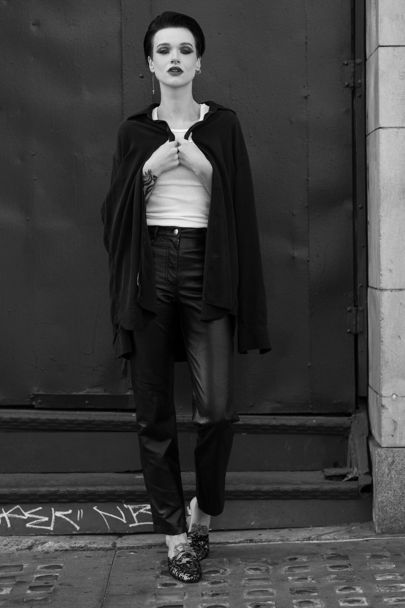 Woman standing in front of a door, wearing a cape, white top, and dark jeans, with dark lipstick and short hair.