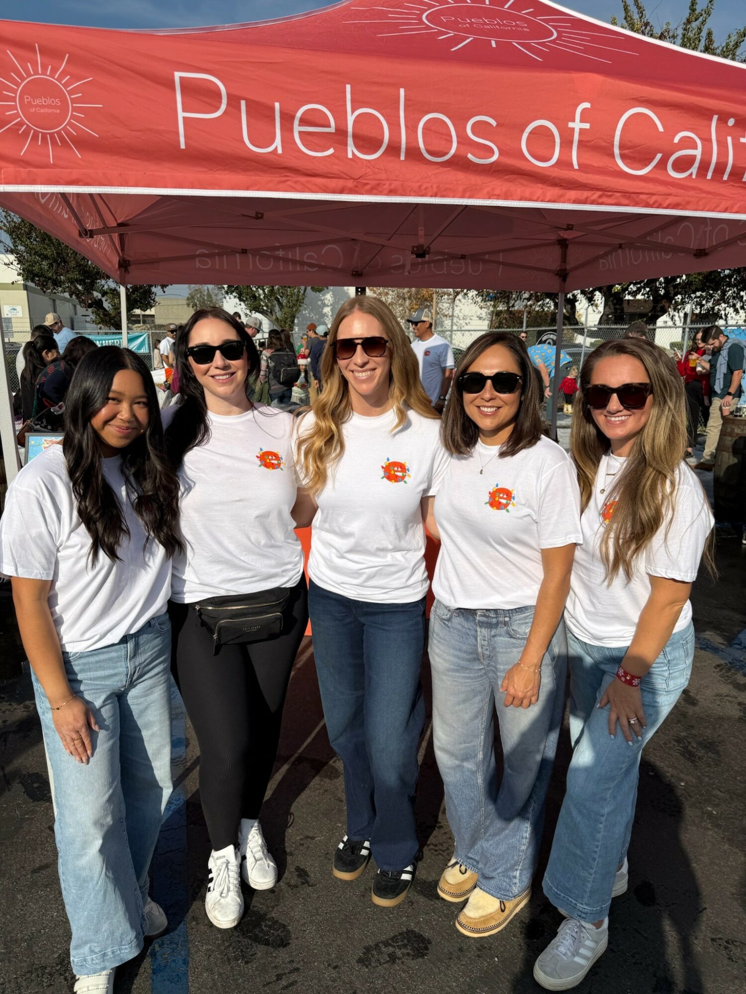 Five women stand outdoors under a red canopy with text, smiling and wearing white shirts and sunglasses.