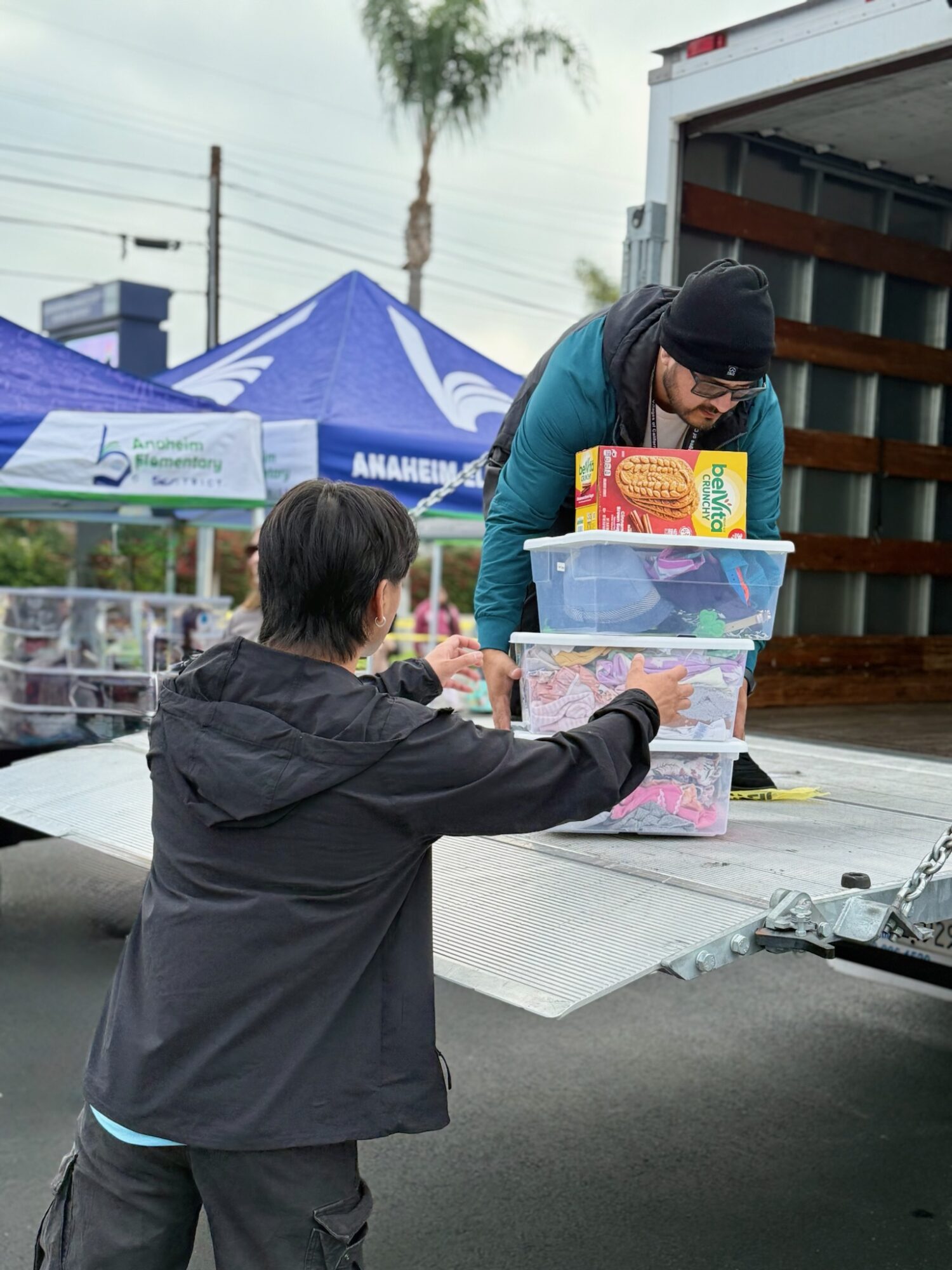 Two people exchanging items at a table outdoors, with tents and a truck in the background.