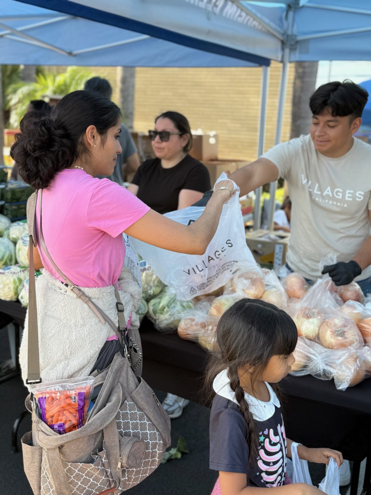 Woman in pink shirt handing a bag of produce to a young man at an outdoor market stall, with a child nearby.