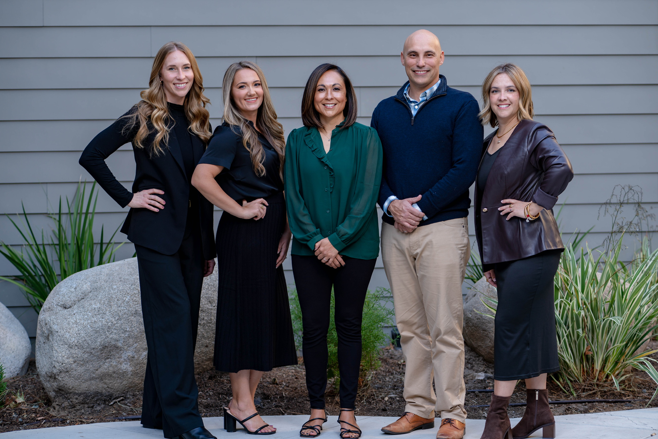 Group of five people standing outdoors in front of a gray wall, smiling, with plants and rocks nearby.