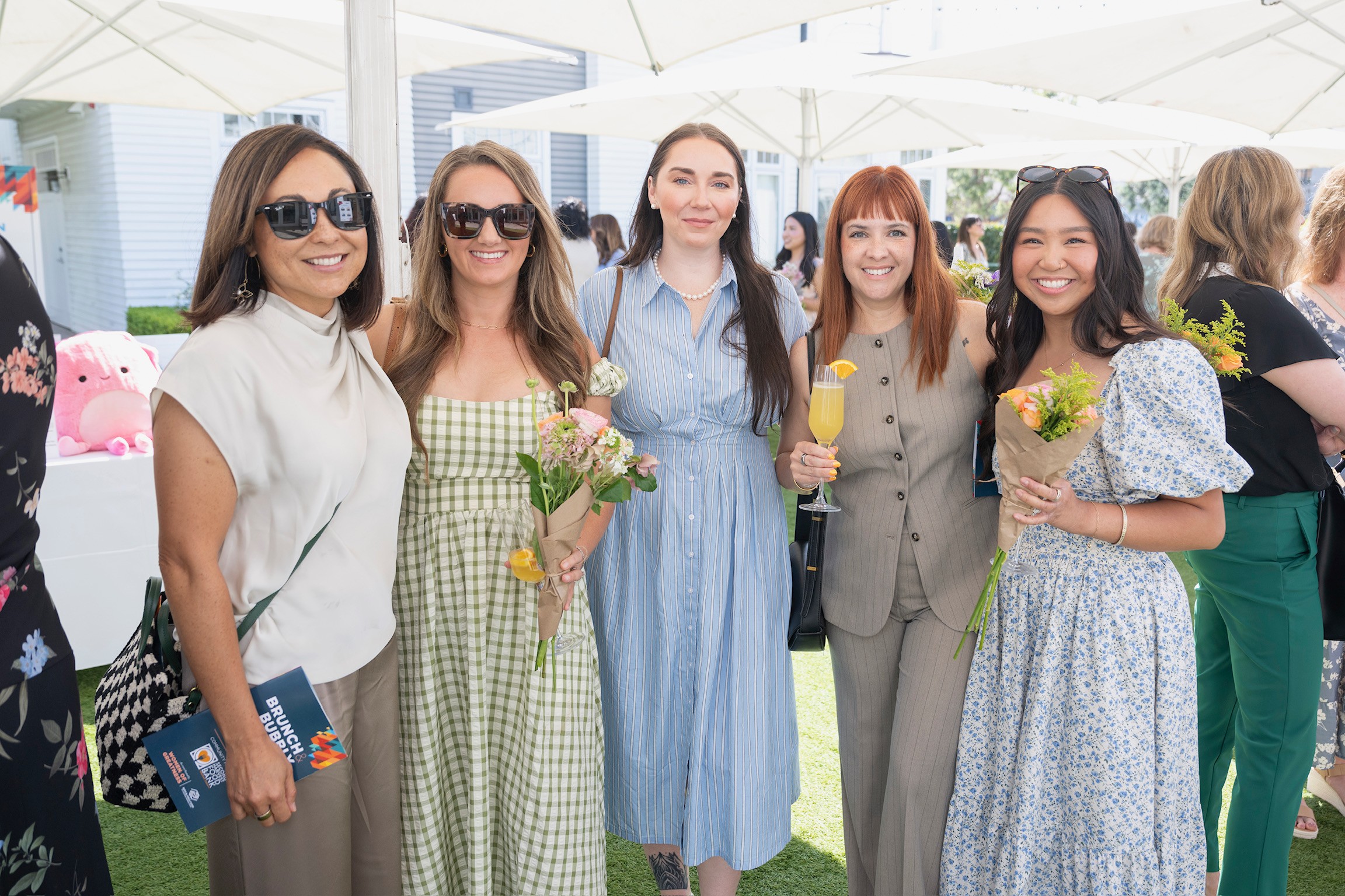 Group of five women smiling outdoors, holding flowers and drinks, under a white canopy, dressed in casual and semi-formal clothing.